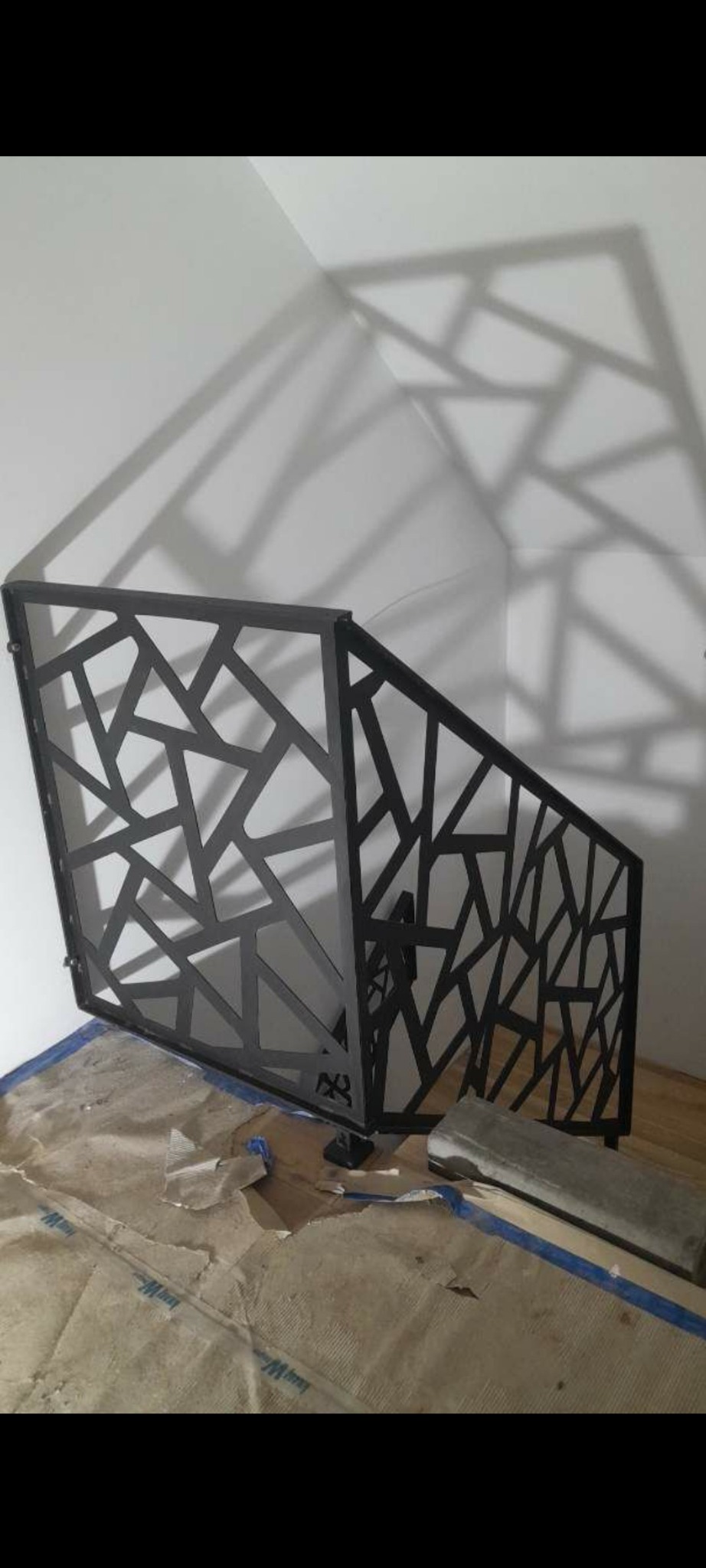 Czarna, metalowa balustrada schodowa o geometrycznym wzorze, w trakcie montażu, zabezpieczona folią podłoga, widoczne narzędzia.