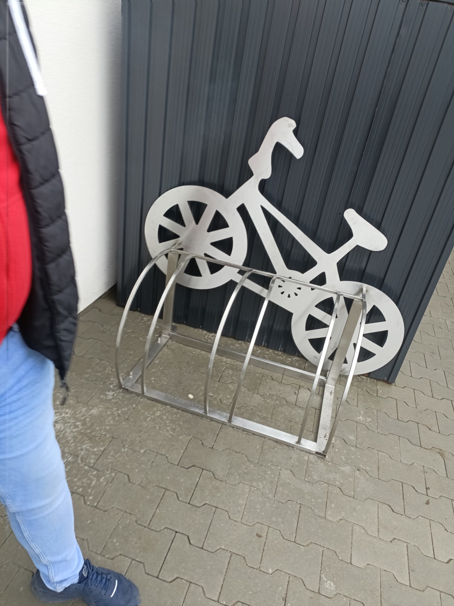 Metalowy stojak na rowery z dekoracyjnym motywem roweru, umieszczony na szarym bruku przed ciemnoszarą ścianą.