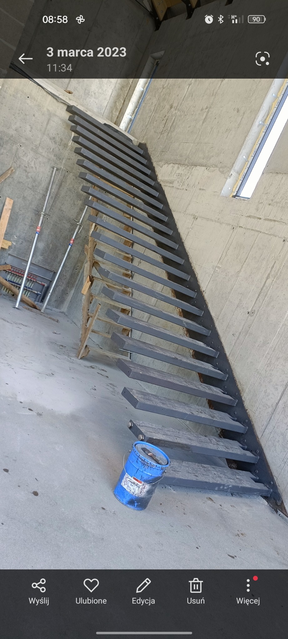 Surowe, metalowe schody w trakcie montażu w surowym wnętrzu, obok stoi niebieska puszka z farbą.