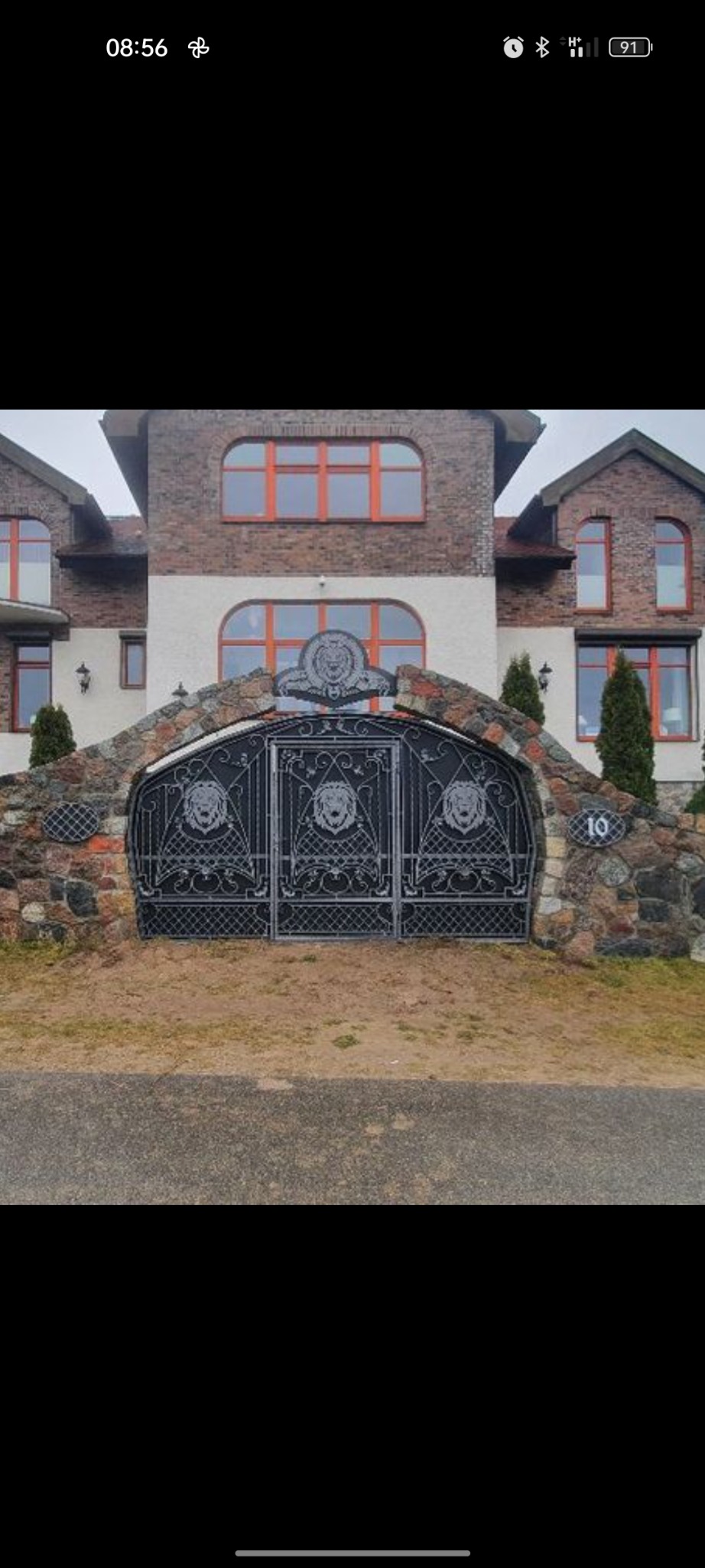 Kuta, dwuskrzydłowa brama wjazdowa z motywem lwa, wkomponowana w kamienny mur, z numerem domu '10', na tle dwupiętrowego domu z cegły i białego tynku.