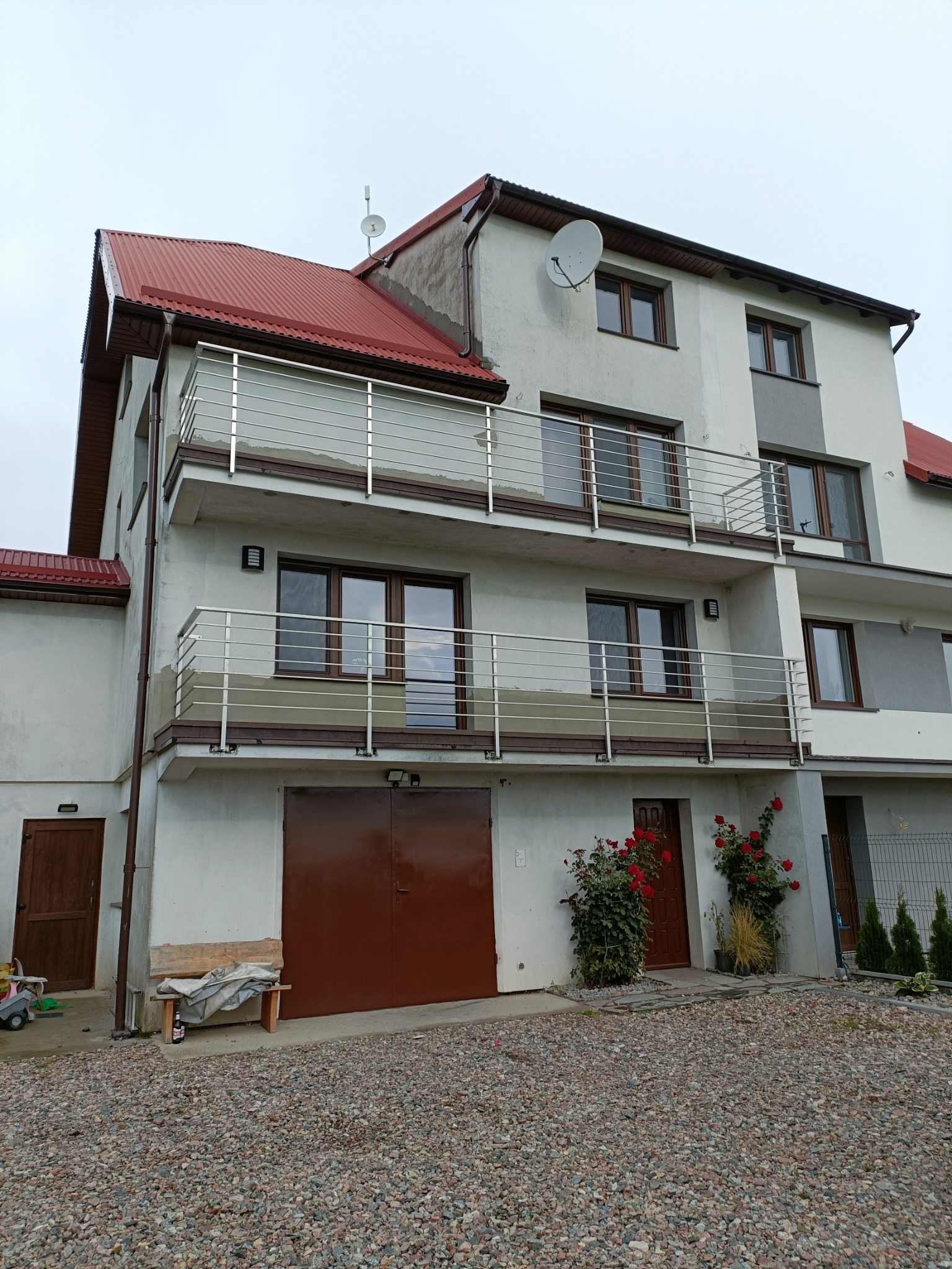 Trzykondygnacyjny dom z balkonami z balustradami ze stali nierdzewnej, czerwonym dachem, brązowymi oknami i drzwiami garażowymi, oraz krzewami róż pnących się po ścianie.