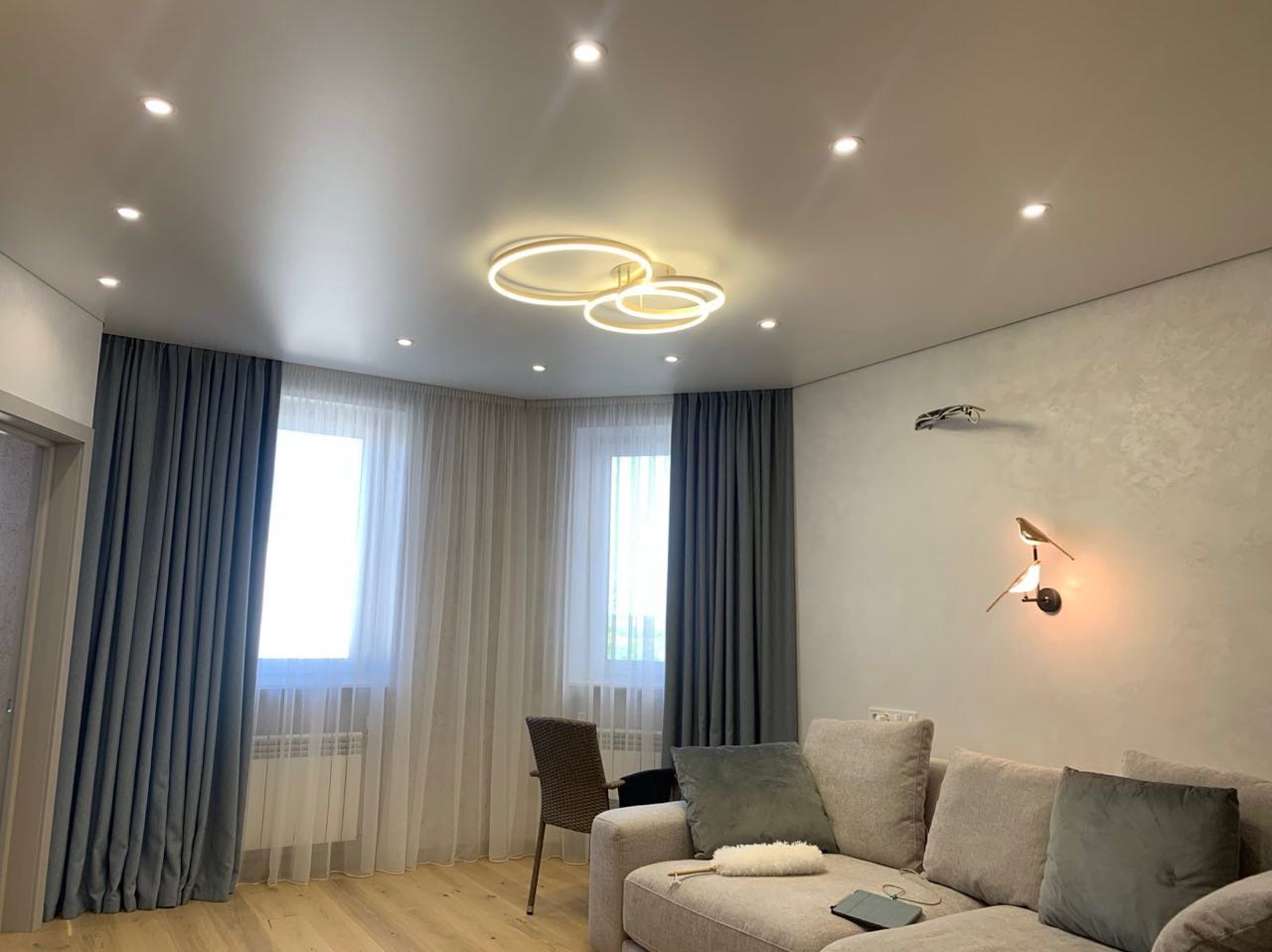 Jasne wnętrze salonu z błyszczącym sufitem, oświetleniem punktowym i lampą wiszącą w kształcie trzech okręgów, szare zasłony i firany przy oknach, beżowa sofa z poduszkami i lampa ścienna...