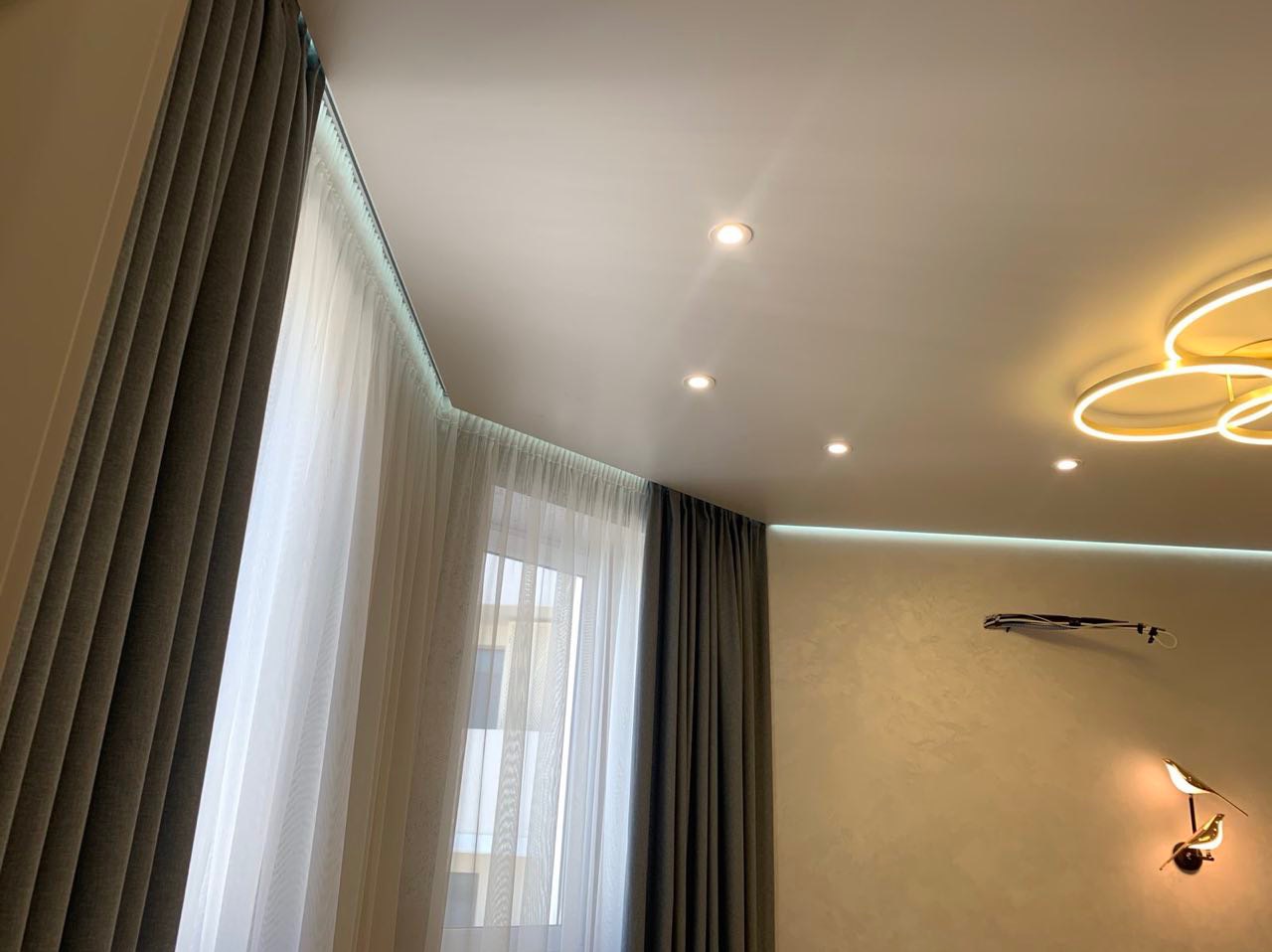 Nowoczesne wnętrze z białym sufitem podwieszanym z oświetleniem punktowym i LED, szare zasłony, firany oraz dekoracyjna lampa ścienna w kształcie ptaków.