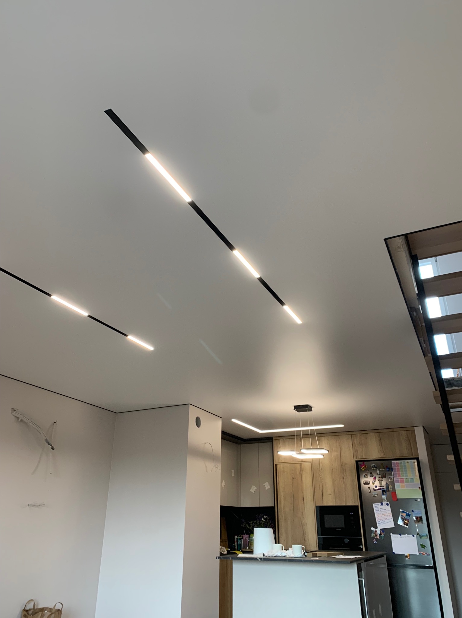 Biały sufit z zamontowanymi czarnymi, liniowymi lampami LED, widok na nowoczesną kuchnię z drewnianymi frontami i schody w tle