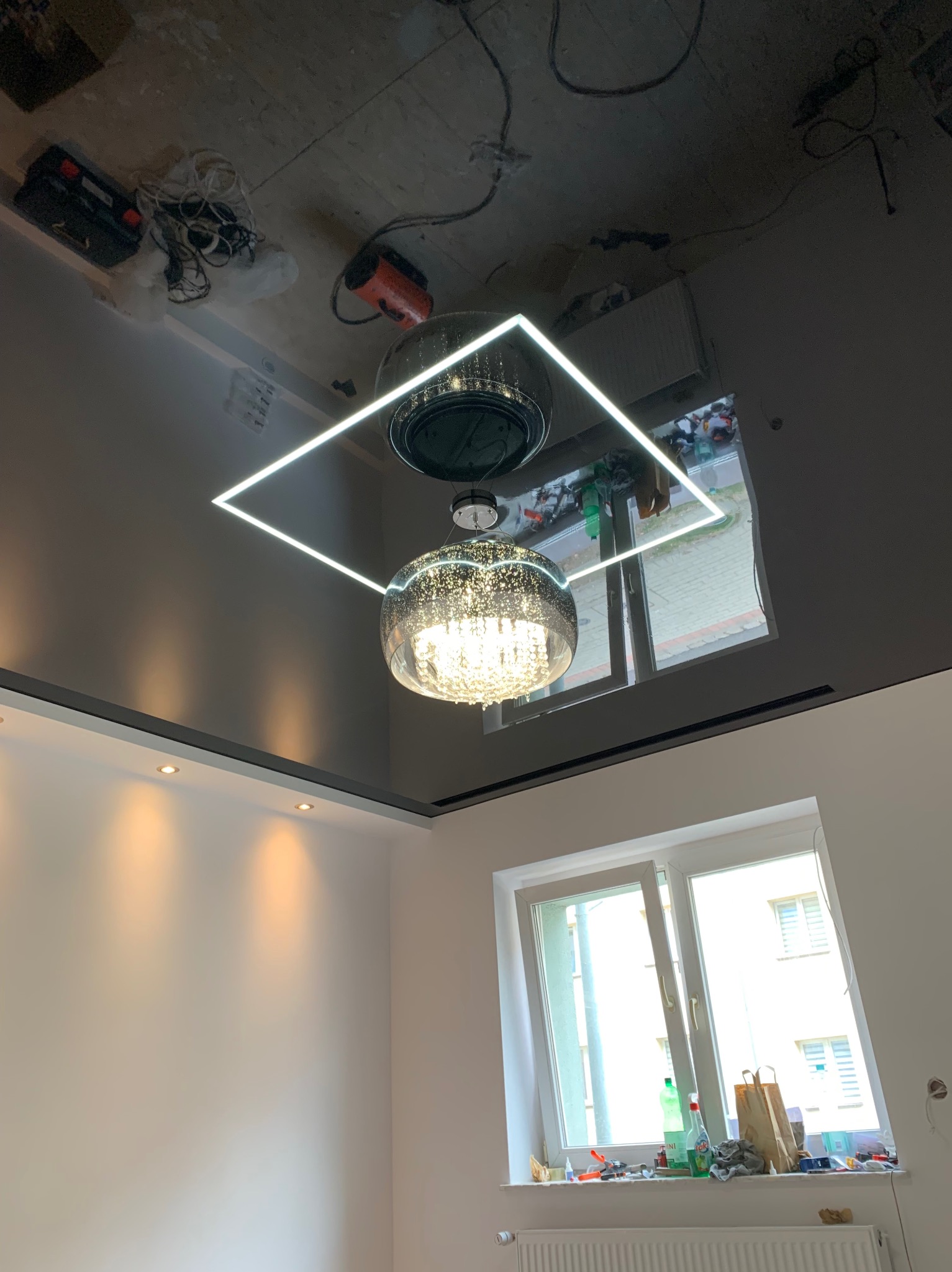 Błyszczący czarny sufit napinany z nowoczesnym oświetleniem: kwadratowa oprawa LED i kryształowy żyrandol, widoczne odbicie okna i narzędzi na suficie.