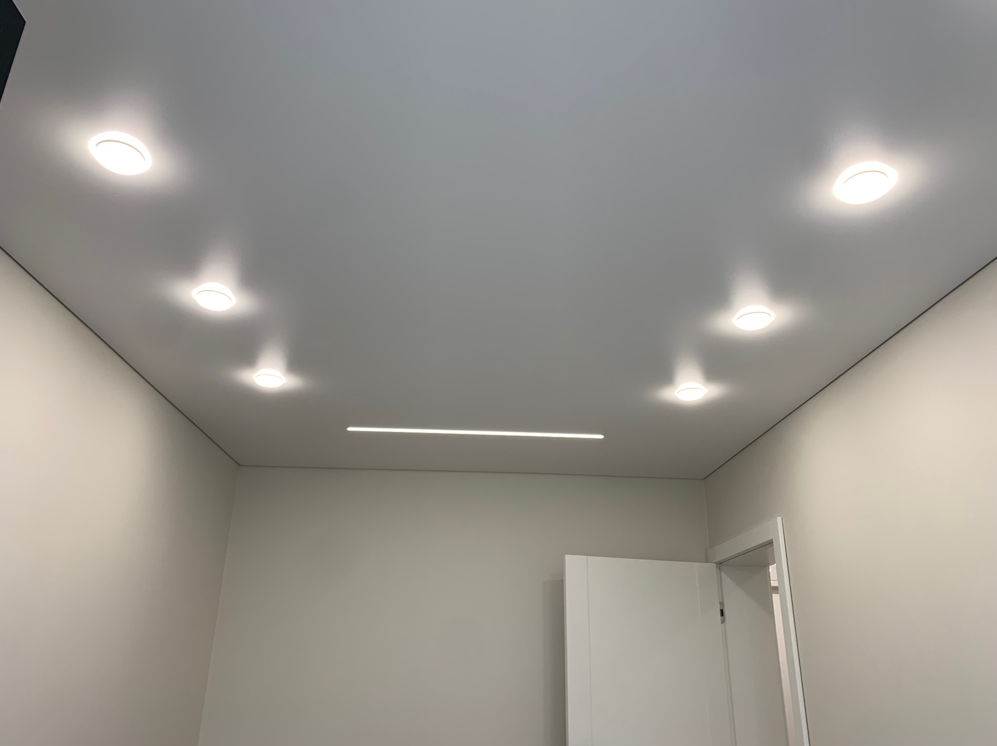 Gładki, szary sufit z sześcioma okrągłymi lampami punktowymi i jedną podłużną lampą liniową, widok z dołu w nowym pomieszczeniu z białymi ścianami i otwartymi drzwiami.