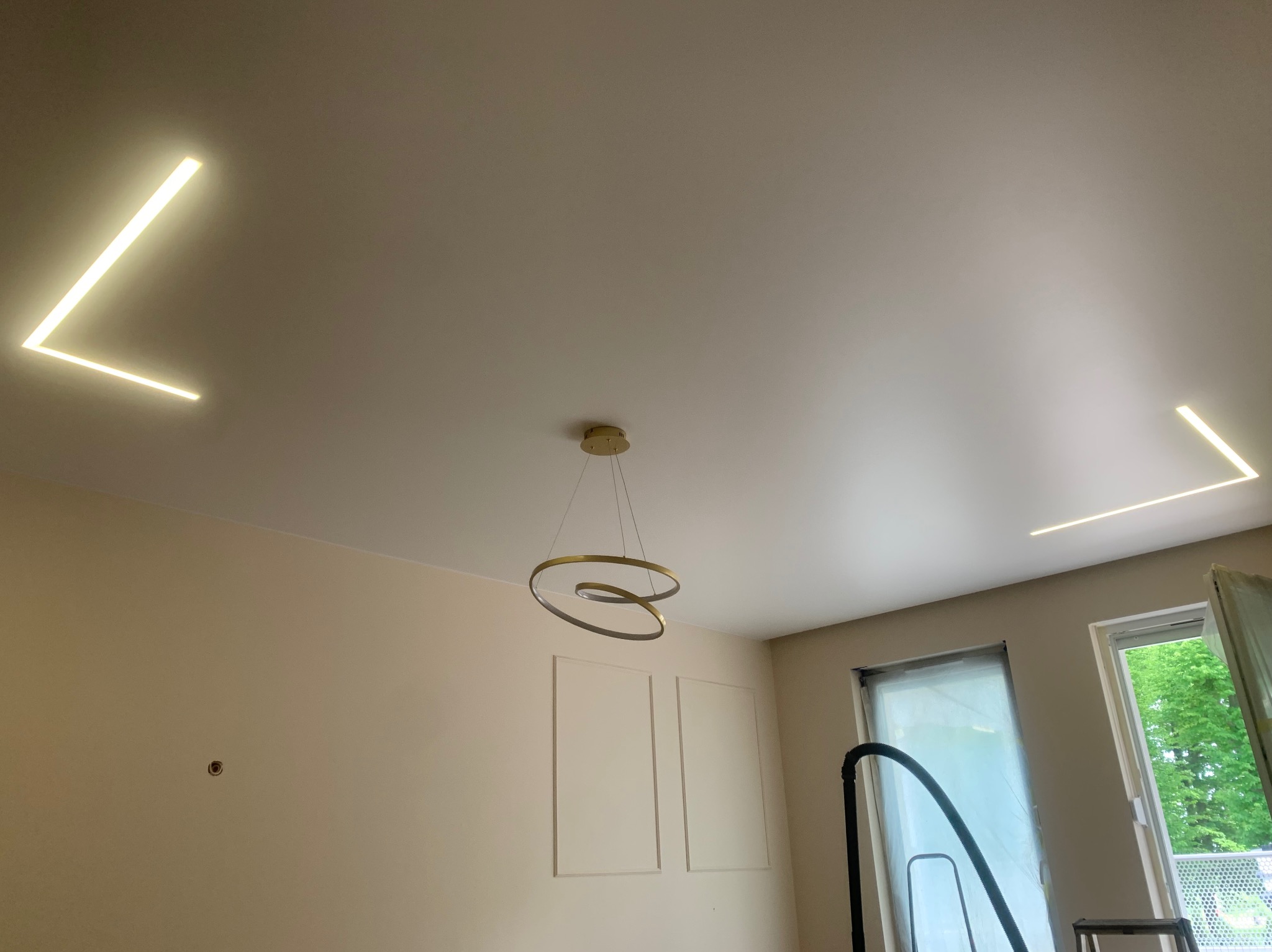 Nowoczesny pokój z białym napinanym sufitem, oświetleniem LED w kształcie litery L w rogach oraz okrągłą, złotą lampą wiszącą na środku.