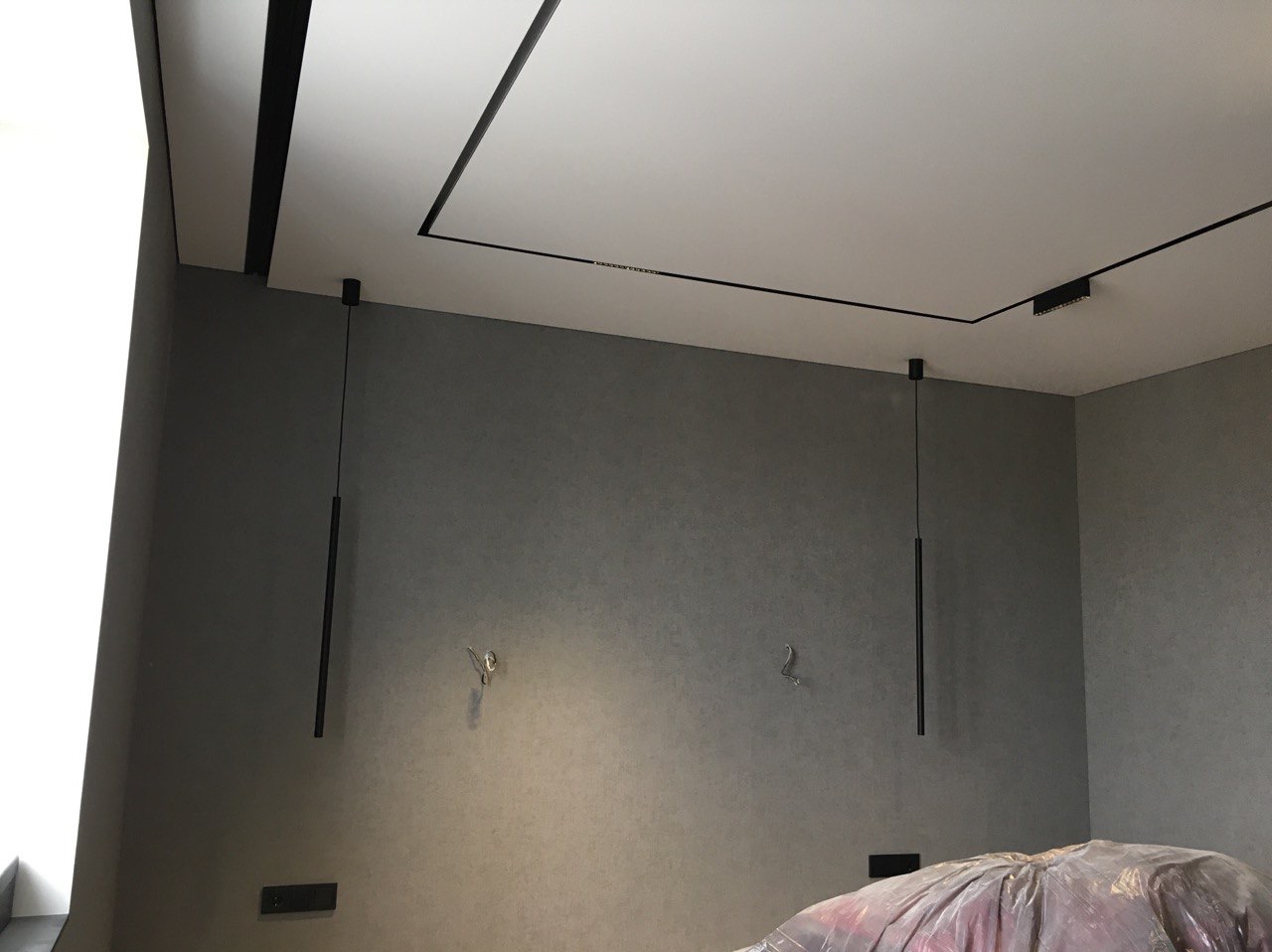 Nowoczesny pokój z szarymi ścianami i białym sufitem z czarnymi liniami LED oraz dwoma podłużnymi lampami wiszącymi na cienkich kablach, częściowo zakryty folią ochronną.