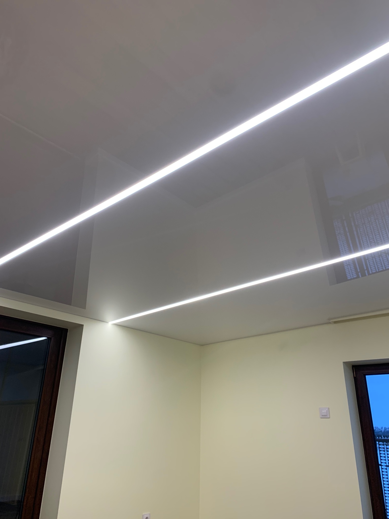 Błyszczący, szary sufit z dwiema liniami wpuszczonego oświetlenia LED, widok z dołu, fragment okna z ciemną ramą w lewym dolnym rogu.