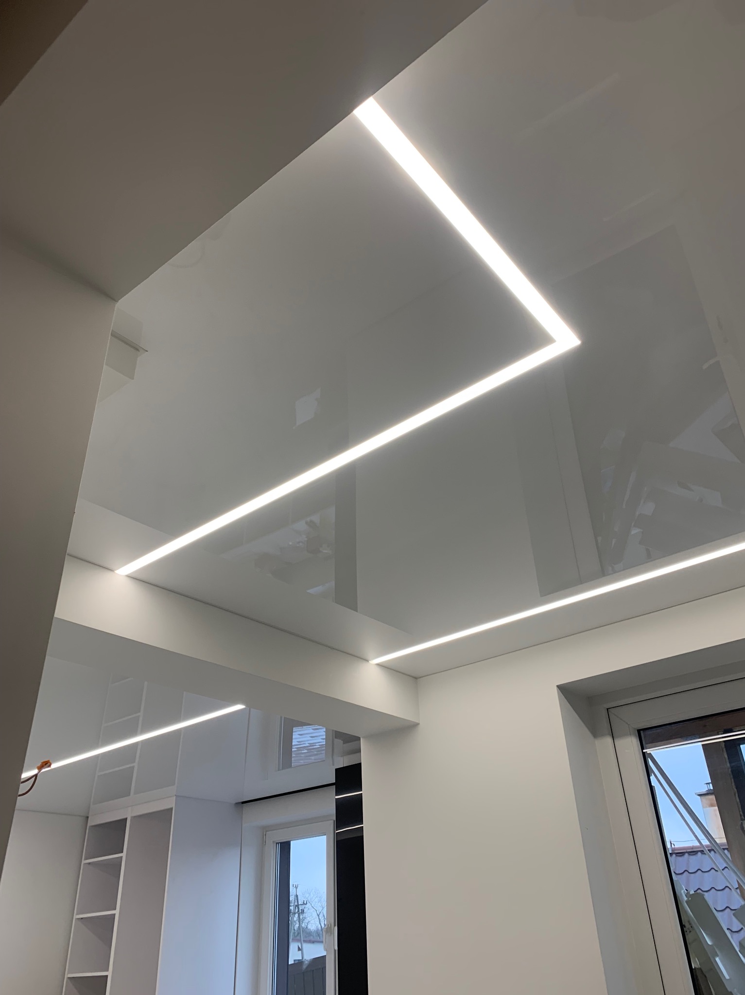 Biały, błyszczący sufit z liniowym oświetleniem LED w kształcie litery L, odbijający światło z okna i wyposażenie pokoju.