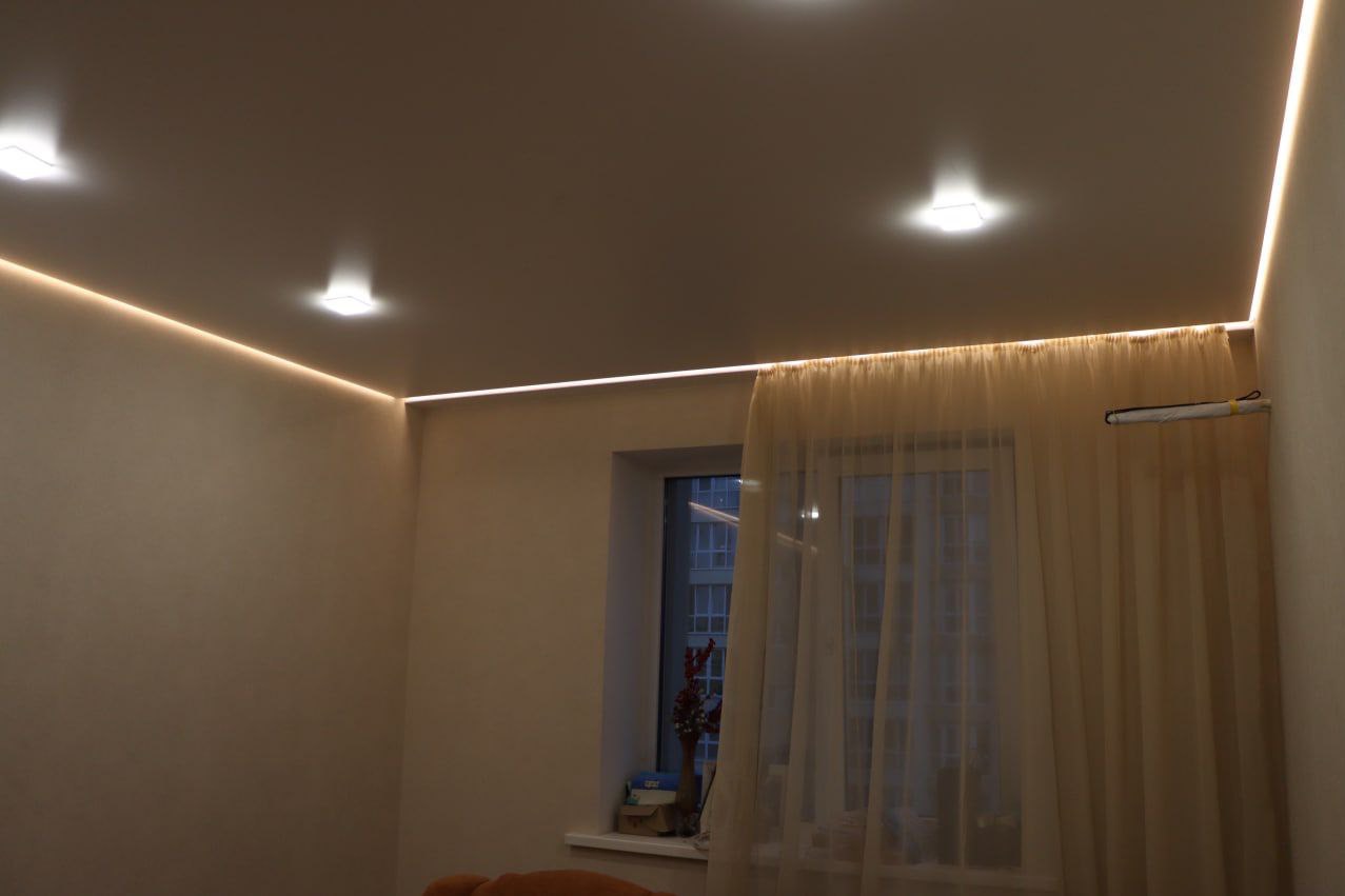 Nowoczesny sufit napinany z oświetleniem LED wzdłuż krawędzi, z reflektorami punktowymi, widok na okno z delikatnymi zasłonami.