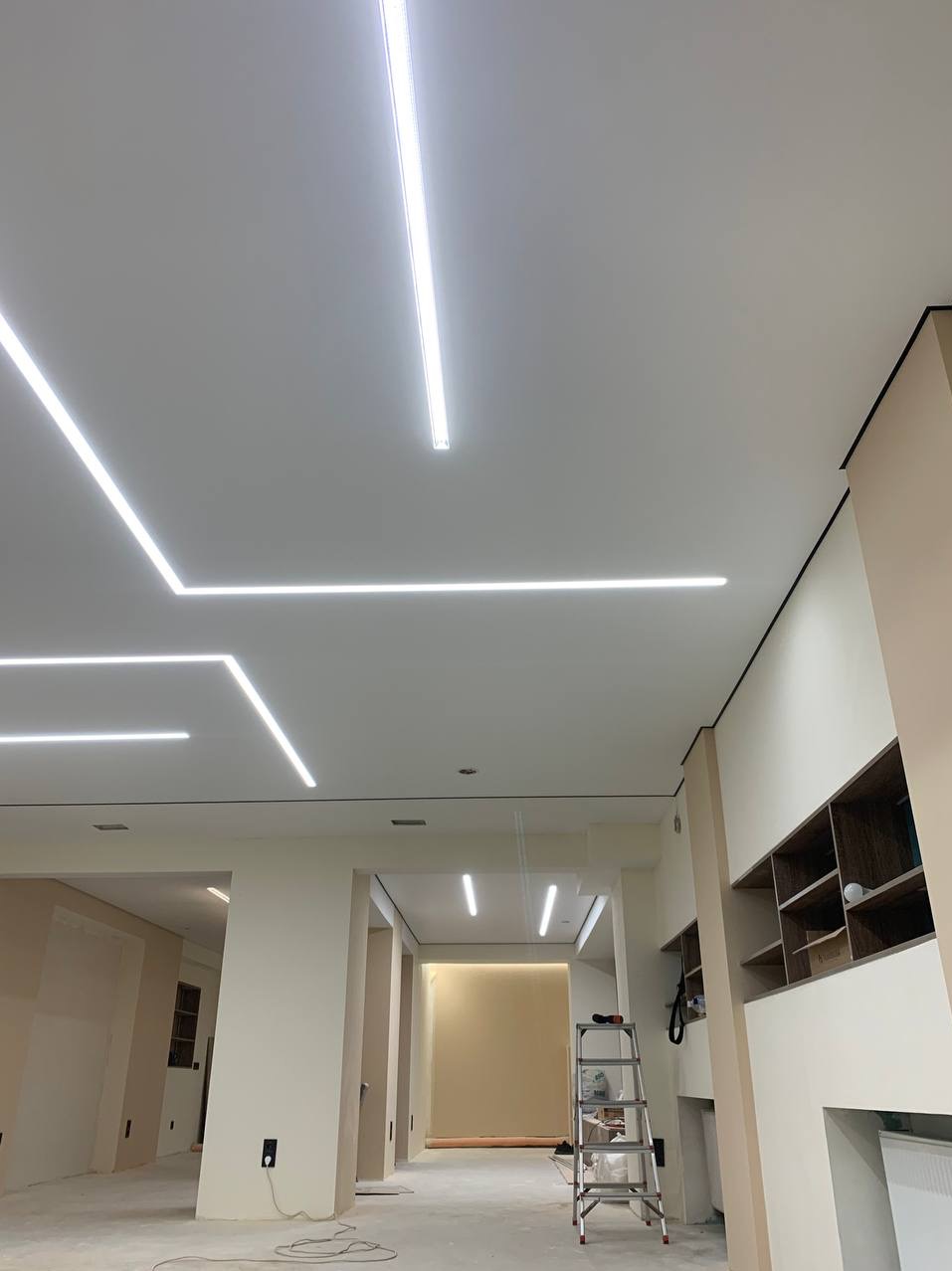Nowoczesne oświetlenie liniowe LED w kształcie zygzaka na białym suficie, widok na wnętrze w trakcie wykańczania, drabina w tle.