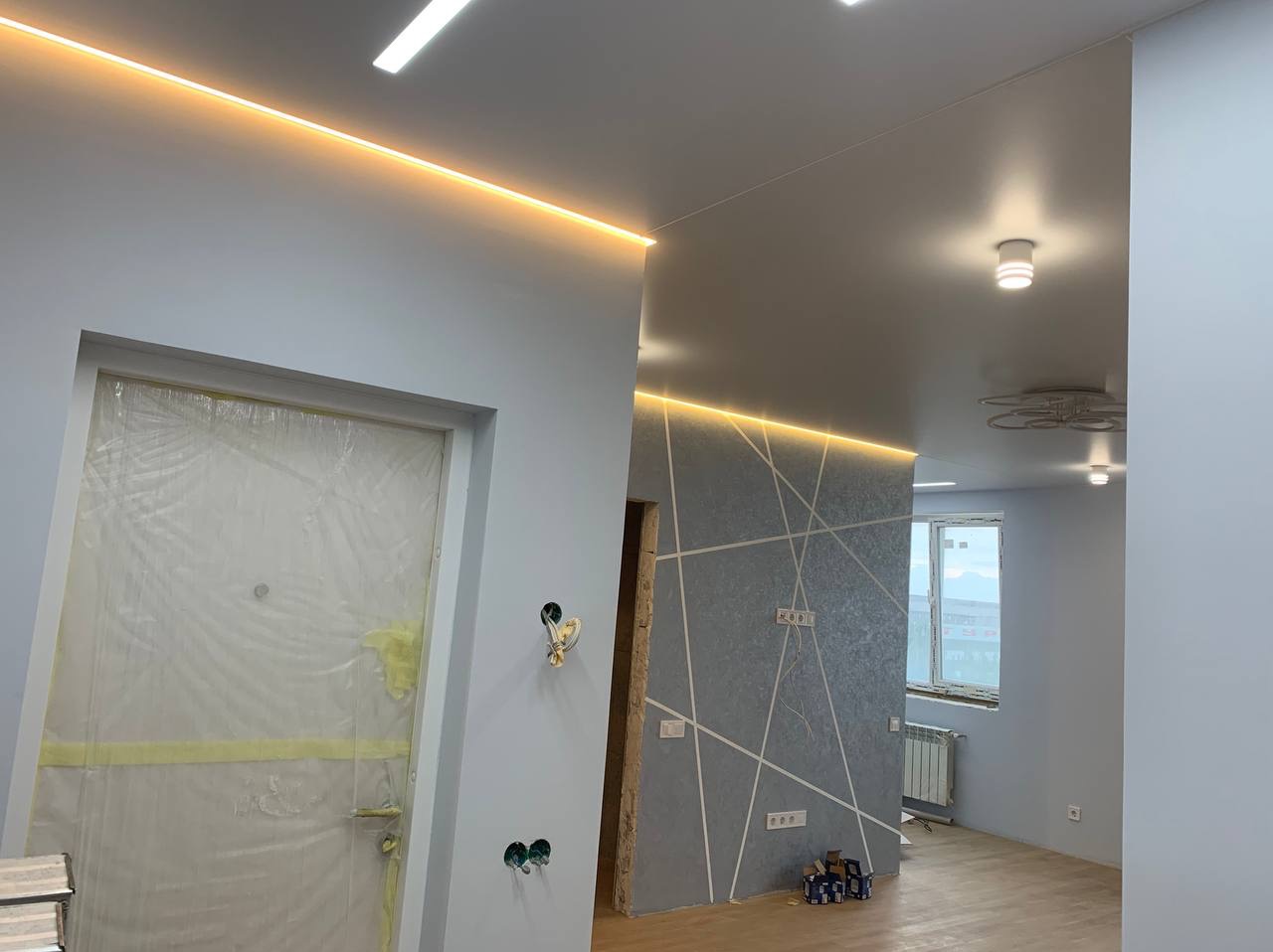 Wnętrze w trakcie remontu: zabezpieczone folią drzwi, ściana z dekoracyjnym wzorem geometrycznym i podświetleniem LED pod sufitem, widoczne puszki elektryczne i grzejnik.