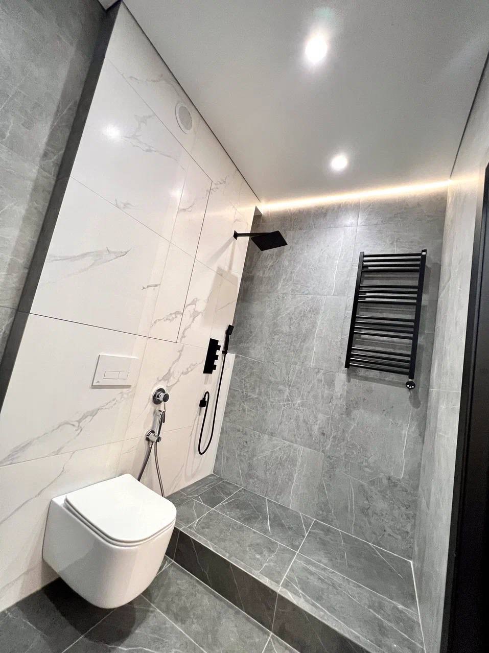 Nowoczesna łazienka z białymi i szarymi marmurkowymi płytkami, czarną armaturą prysznicową i grzejnikiem drabinkowym, oświetleniem LED i podwieszaną toaletą.