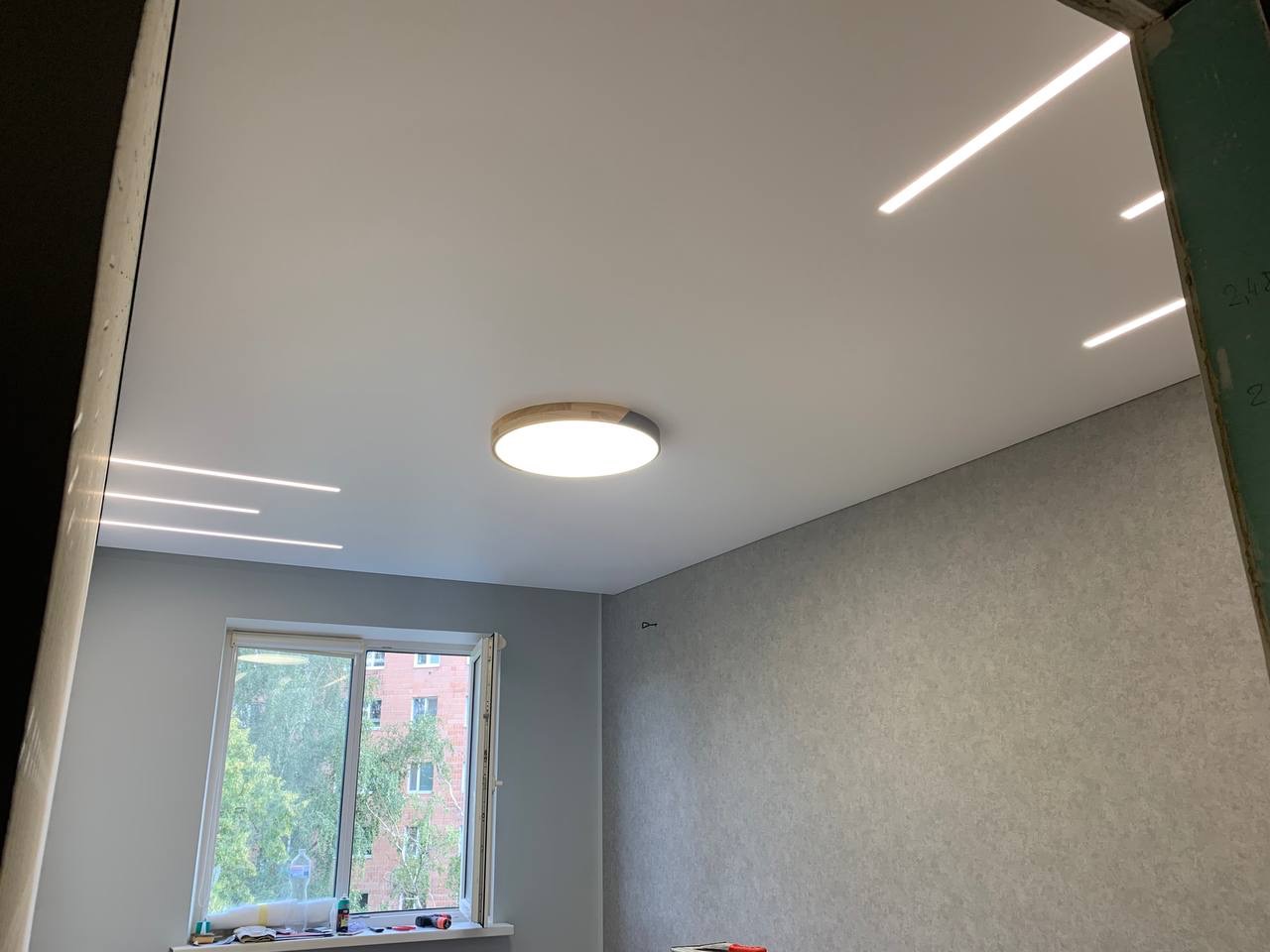 Biały sufit z oświetleniem LED liniowym i okrągłą lampą centralną, widok z dołu na tle okna z widokiem na budynek i zielone drzewa.