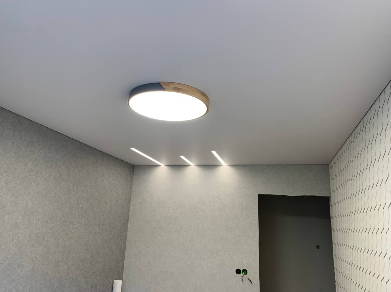 Biały sufit napinany z okrągłą lampą z drewnianym akcentem i trzema liniowymi oprawami oświetleniowymi LED, szare ściany z różnymi fakturami, widoczne gniazdka elektryczne.