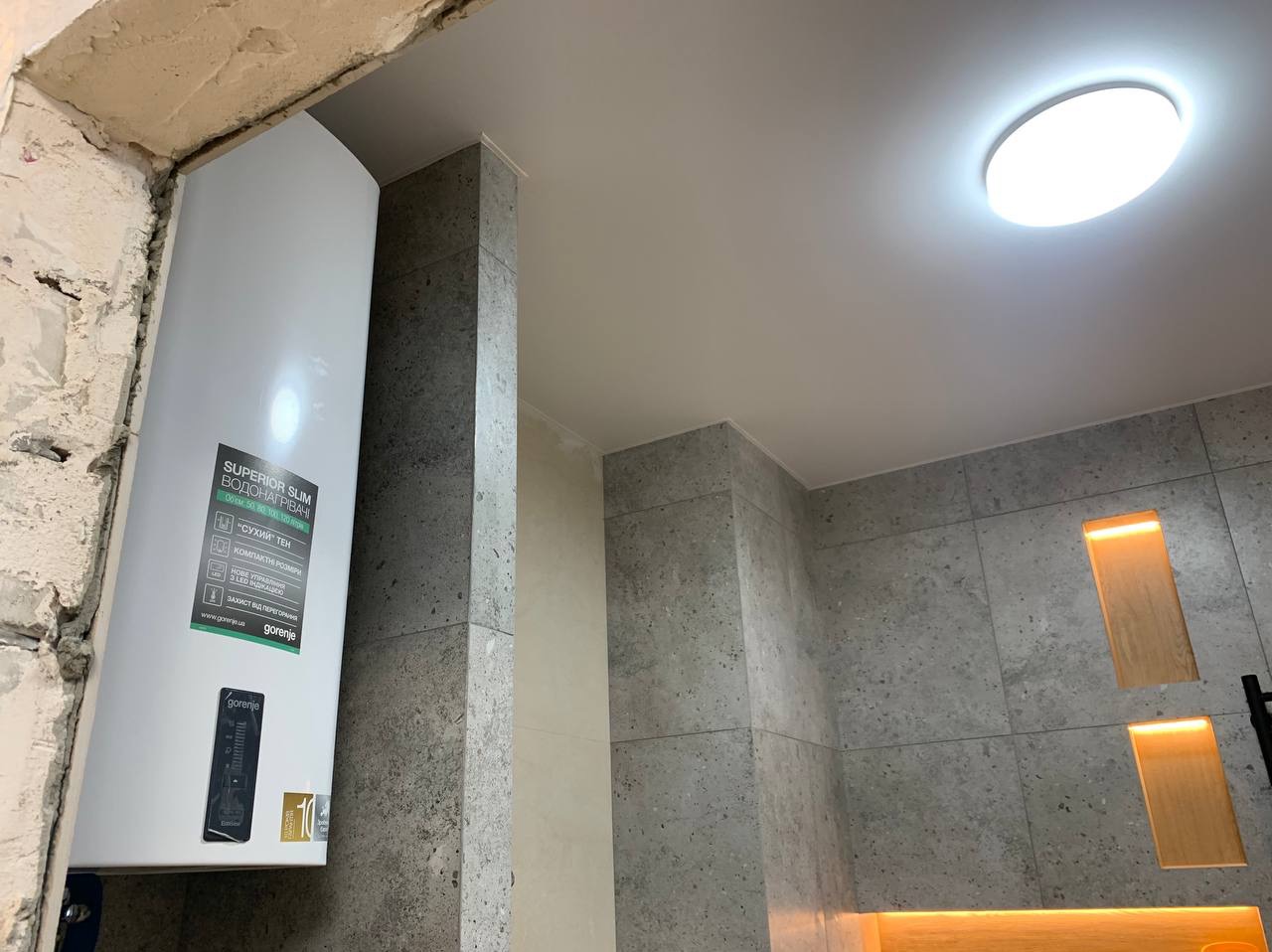 Łazienka po remoncie: szara, kamienna okładzina ścian, podwieszany sufit z okrągłym oświetleniem LED, oraz biały, wąski bojler elektryczny Gorenje zamontowany na ścianie, widoczny fragment surowego...