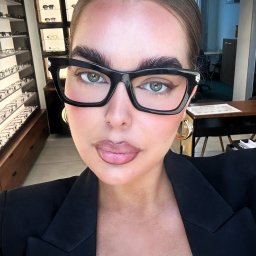 OKOOPTYK - Portret kobiety w czarnych okularach korekcyjnych, z widoczną ekspozycją oprawek w tle, sugerujący wizytę u okulisty.