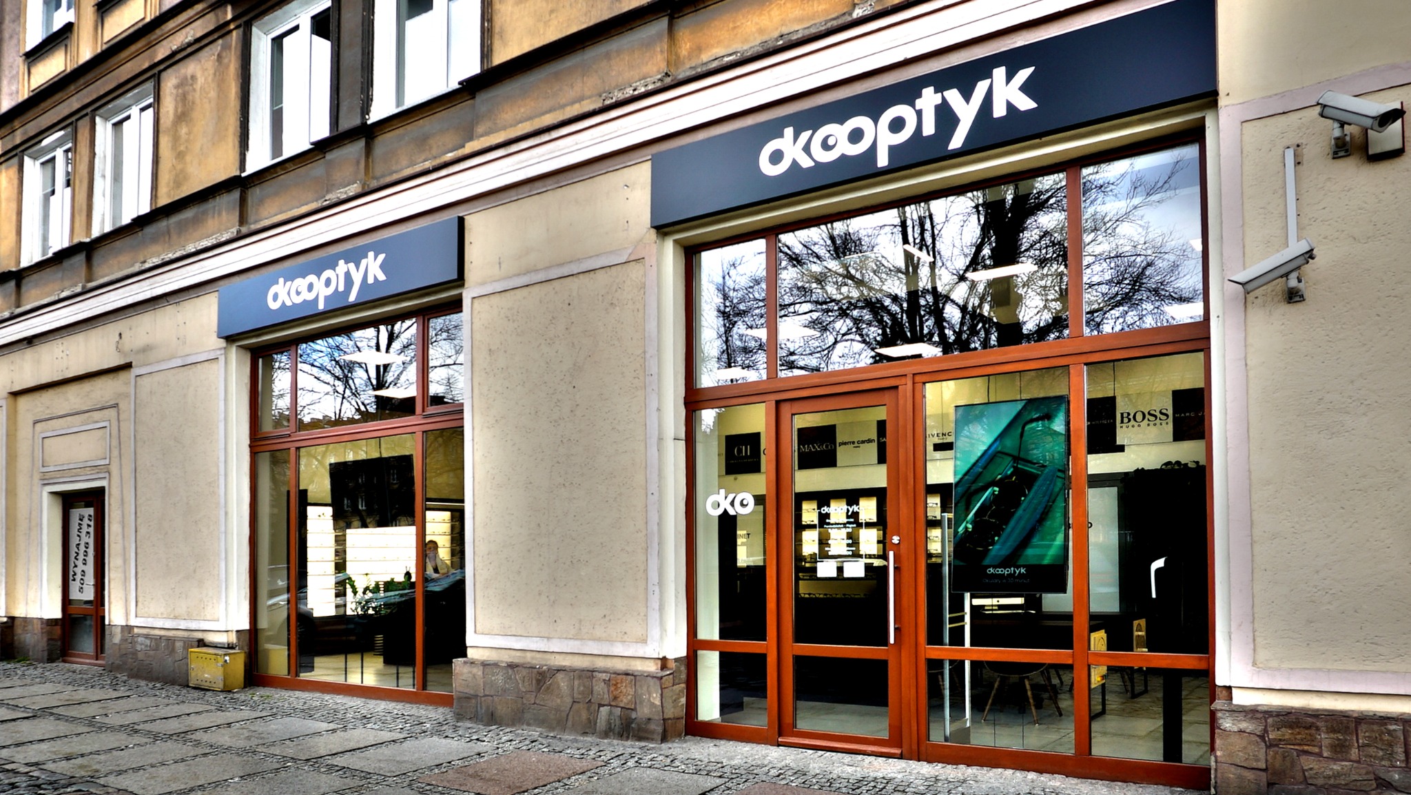 Fasada budynku z dwoma wejściami do salonu optycznego DkOptyk, z szyldami nad wejściami, widoczne kamery monitoringu na elewacji, oraz reklamy okularów w witrynach.