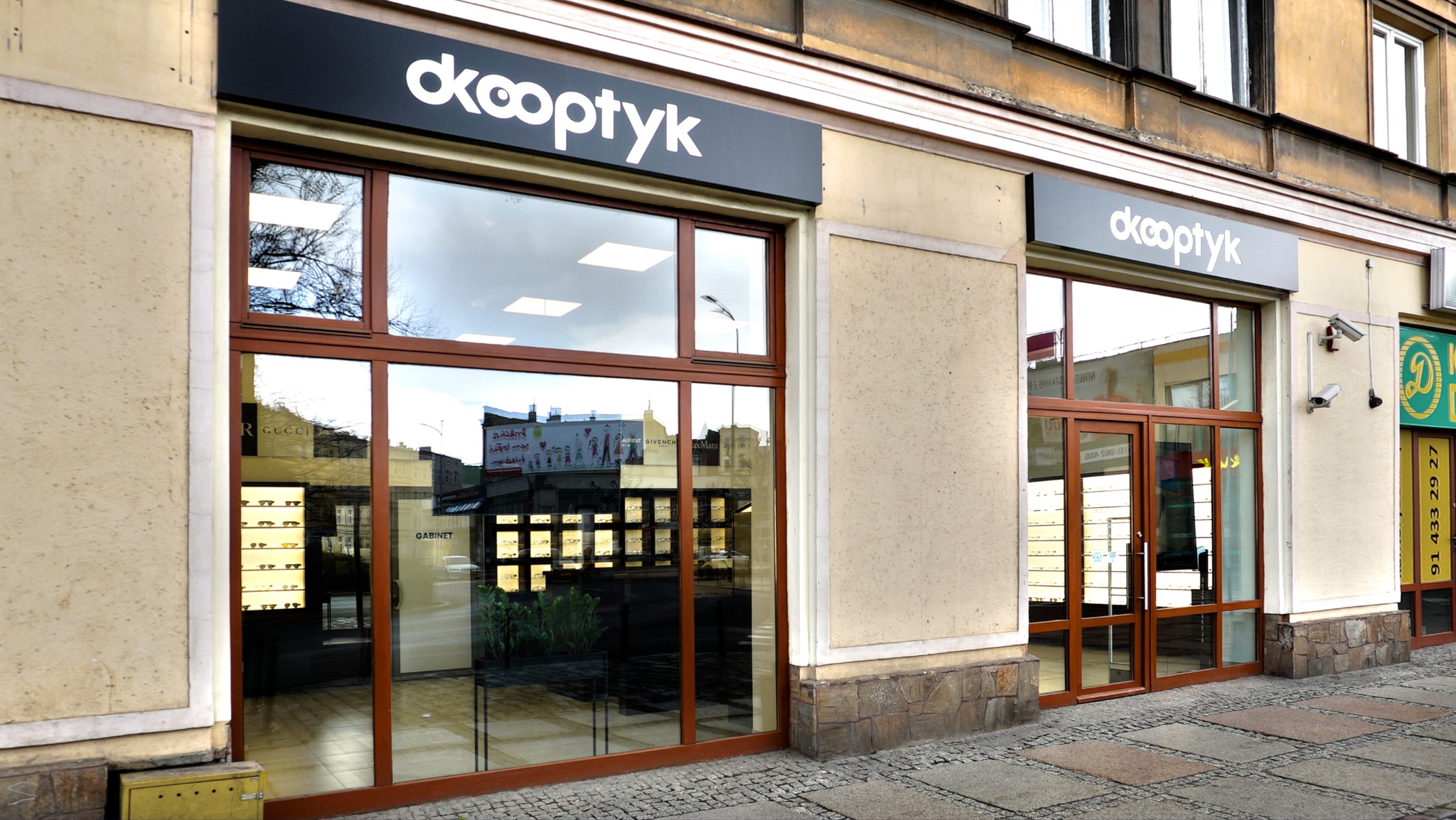 Fasada budynku z dużymi oknami wystawowymi salonu optycznego z szyldem 'dkOoptyk' i widocznymi okularami wewnątrz.