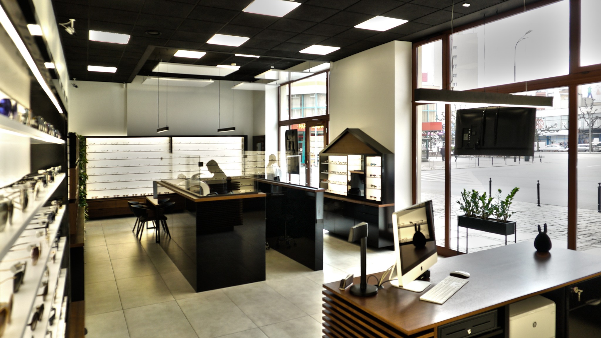 Wnętrze salonu optycznego z ekspozycją oprawek, ciemnymi meblami i widokiem na ulicę przez duże okno.