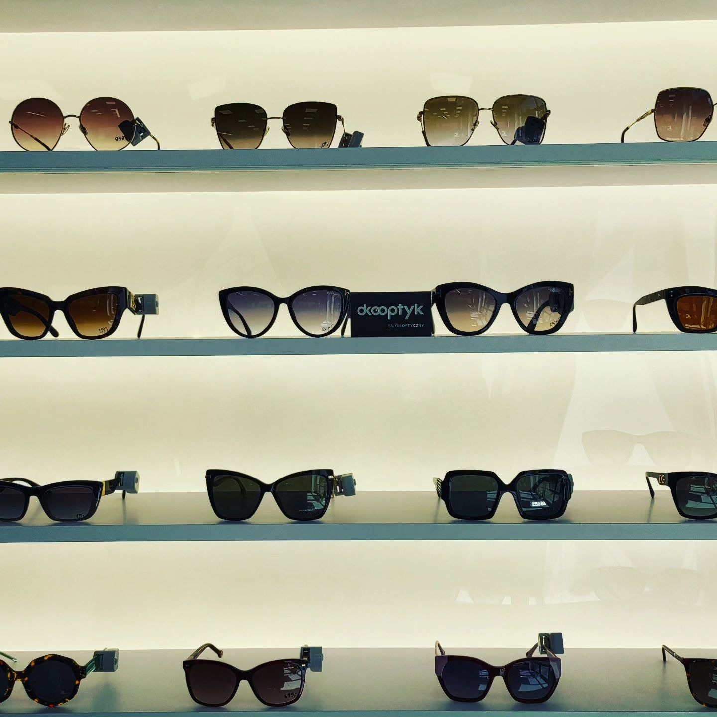Ekspozycja okularów przeciwsłonecznych różnych marek na czterech półkach w salonie optycznym, widoczne metki na niektórych modelach.