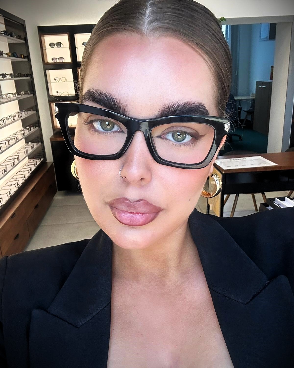 Portret kobiety w czarnych okularach korekcyjnych, z widoczną ekspozycją oprawek w tle, sugerujący wizytę u okulisty.