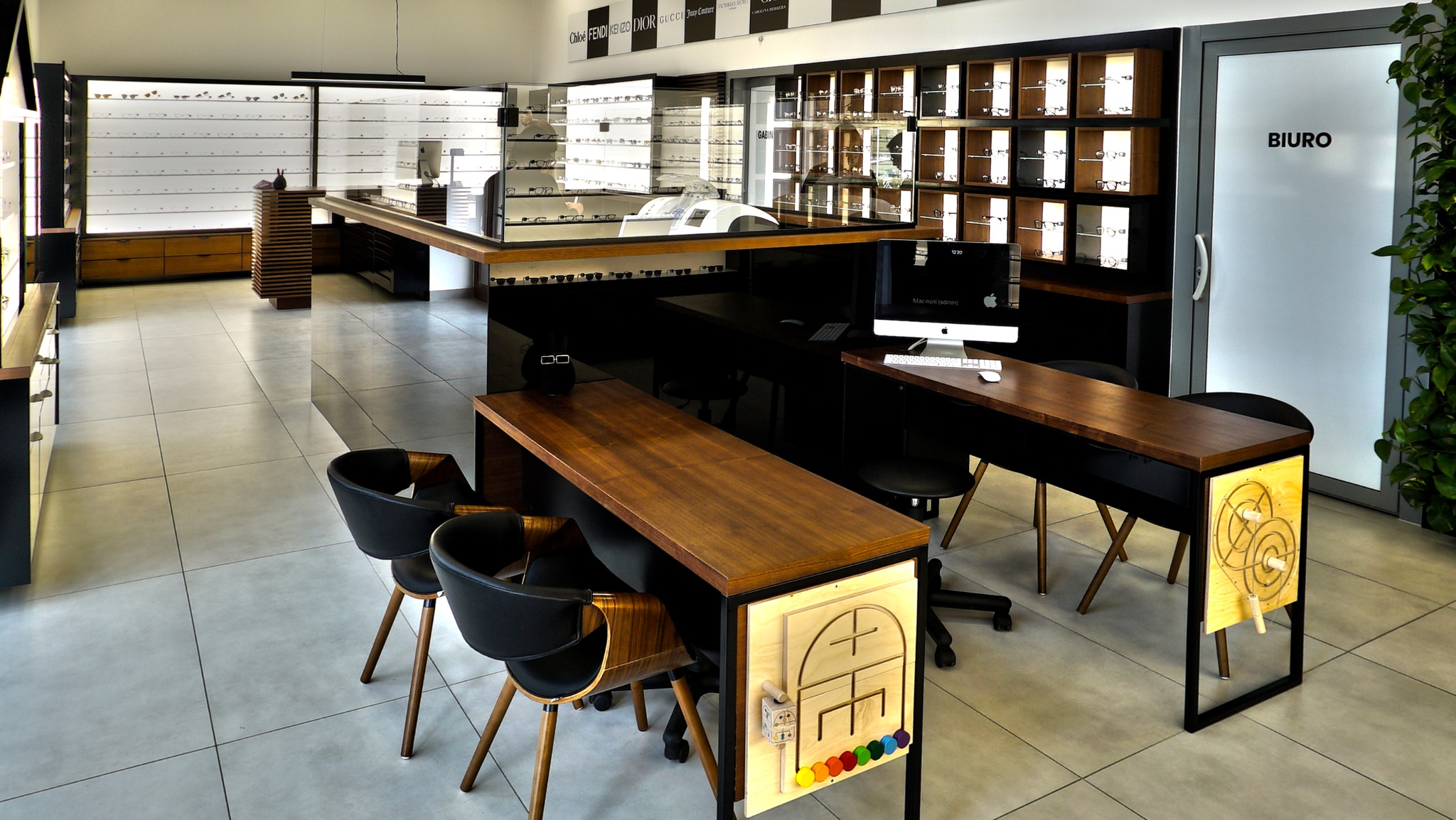 Wnętrze salonu optycznego z ekspozycją okularów w gablotach, biurkami z komputerami Apple oraz drewnianymi elementami wykończenia. Na pierwszym planie stolik z edukacyjną zabawką dla dzieci.