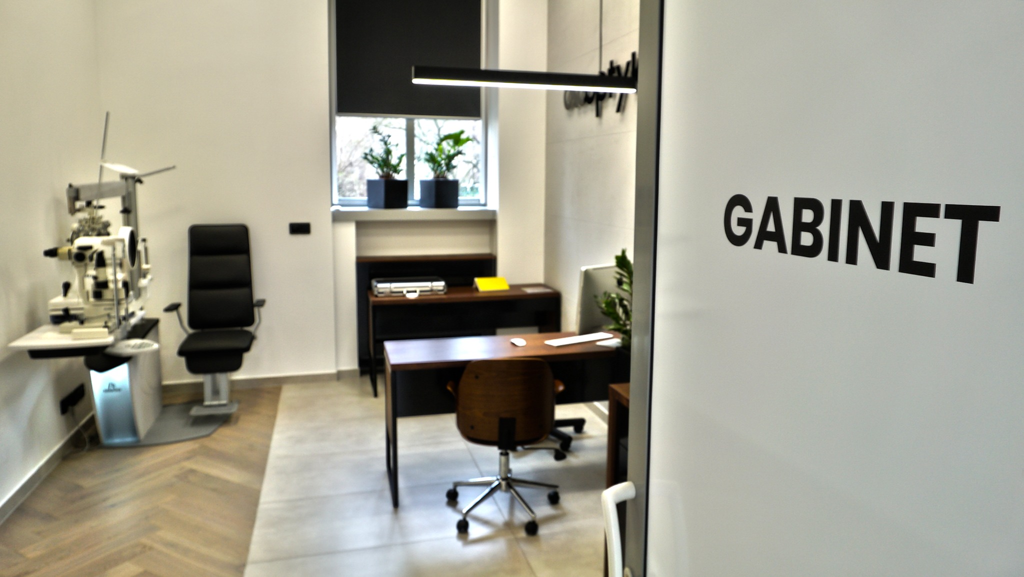 Wnętrze gabinetu lekarskiego z aparaturą diagnostyczną, biurkiem i krzesłem, widoczne przez uchylone drzwi z napisem 'GABINET'.