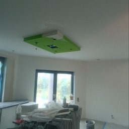 ROMAN SAVCHYSHYN - Narożny sufit podwieszany z zielonym akcentem w surowym wnętrzu. Widoczne narzędzia malarskie i zabezpieczenia podłogi folią, okno w tle.