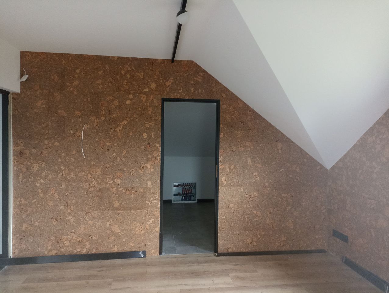 Pokój na poddaszu z wykończeniem ścian korkiem. Widoczne wejście do innego pomieszczenia, oświetlenie sufitowe i skośny sufit.