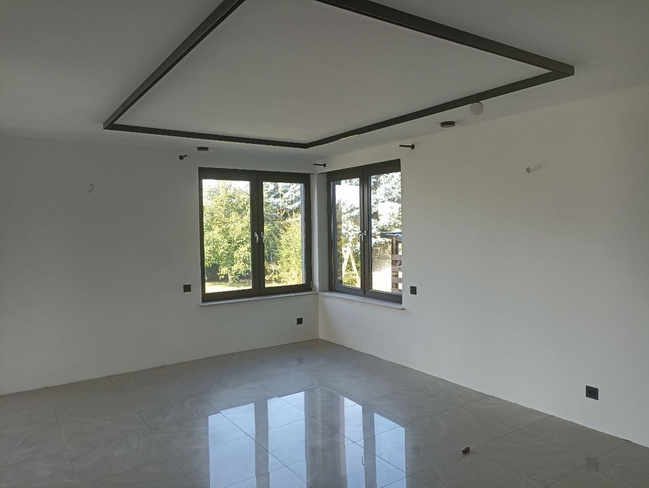 Puste, jasne wnętrze z szarą ramą na suficie, dwoma oknami z widokiem na zieleń i błyszczącą podłogą z płytek. Gładkie, białe ściany i minimalistyczny design.