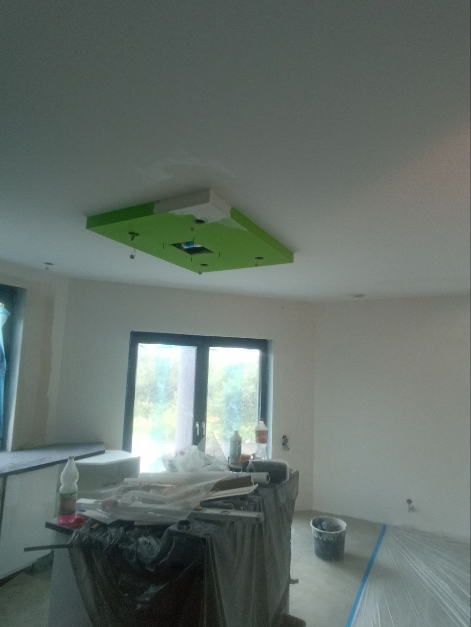 Narożny sufit podwieszany z zielonym akcentem w surowym wnętrzu. Widoczne narzędzia malarskie i zabezpieczenia podłogi folią, okno w tle.