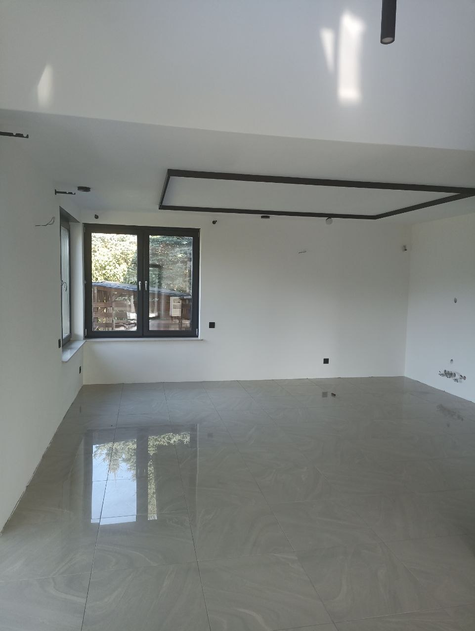 Nowoczesne wnętrze z szarymi płytkami podłogowymi, dużym oknem z widokiem na zieleń i minimalistycznym oświetleniem sufitowym.