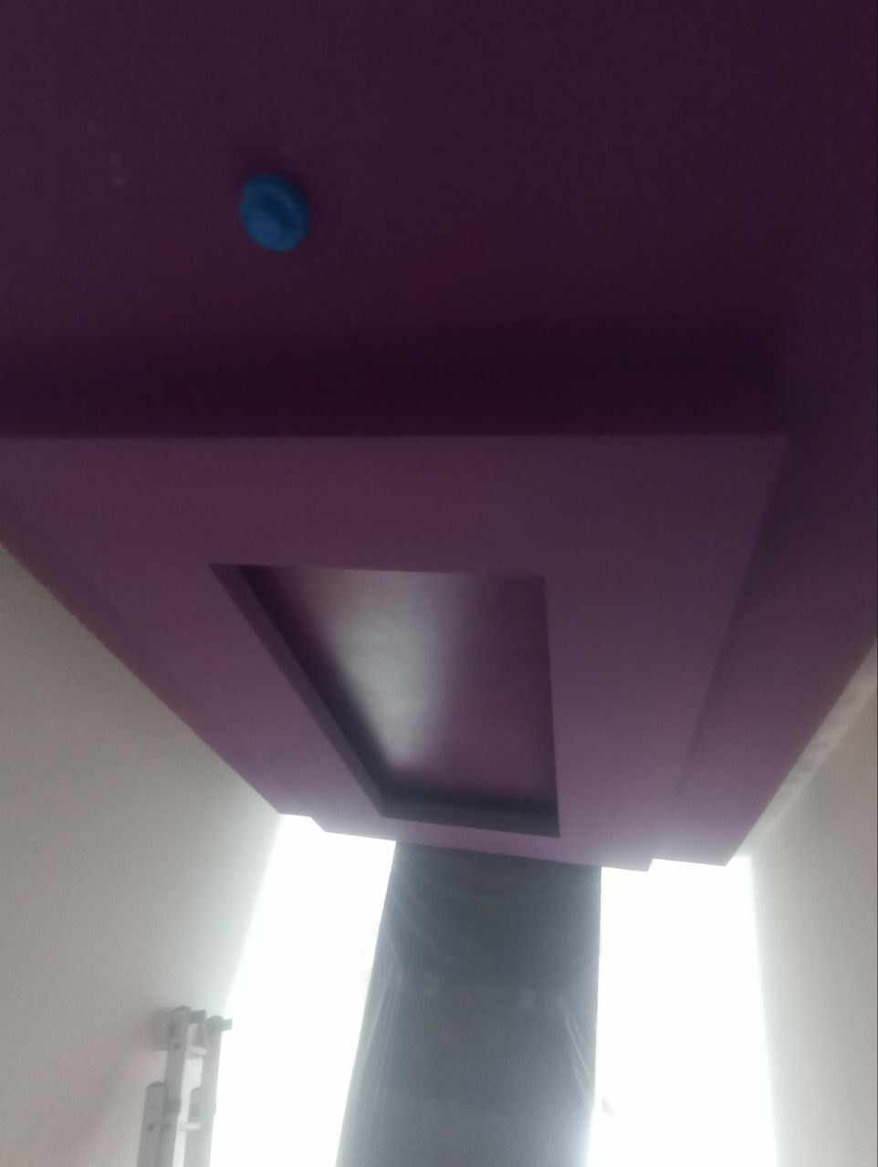 Fioletowy sufit podwieszany z centralnym wgłębieniem, zabezpieczony niebieską zaślepką, widoczny z dołu pod kątem, z fragmentem drabiny malarskiej.