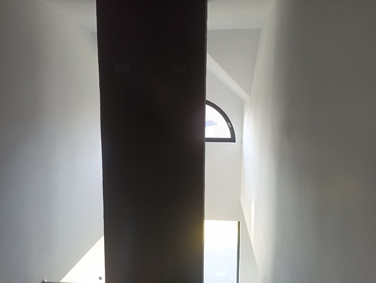 Wnętrze klatki schodowej z oknem w kształcie półkola, ciemnym filarem po lewej i białymi ścianami. Ujęcie od dołu, z widocznym oświetleniem.