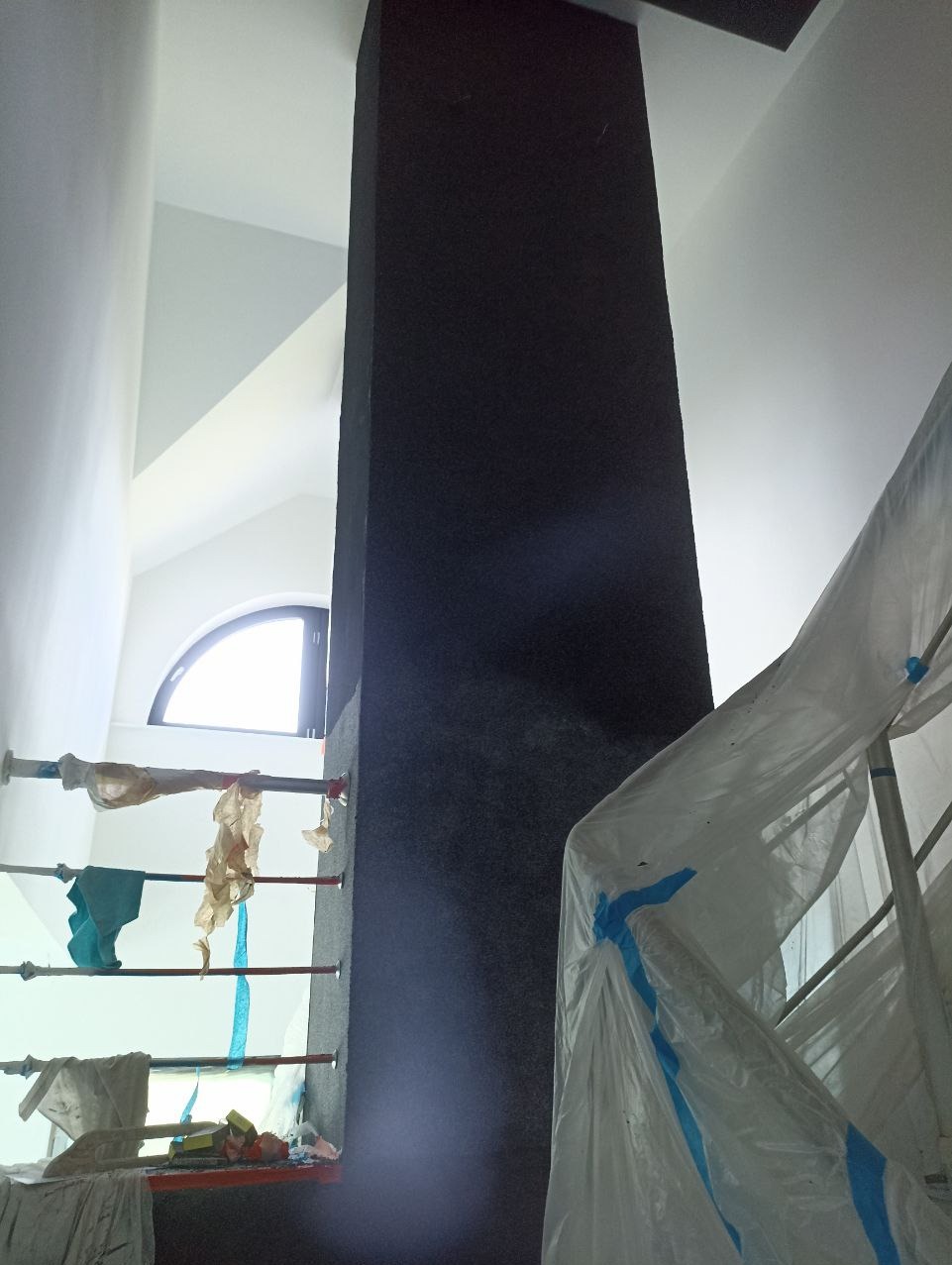 Wnętrze w trakcie prac wykończeniowych: pionowy element konstrukcyjny w ciemnym kolorze, zabezpieczony folią rusztowania, okno łukowe w tle.