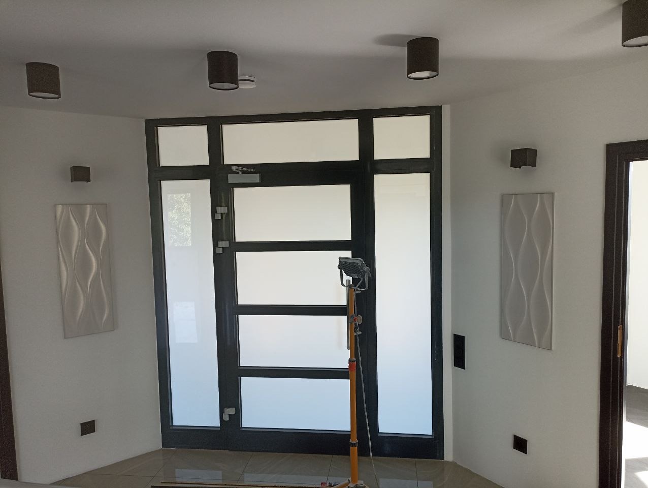 Nowoczesne wnętrze z oświetleniem punktowym i czarną stolarką okienną. Na ścianach dekoracyjne panele 3D. Widoczny statyw oświetleniowy w trakcie montażu.