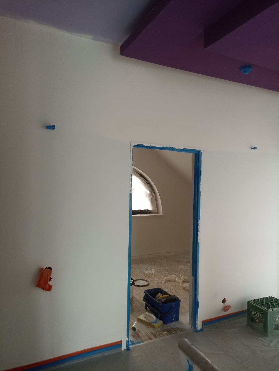 Białe ściany po malowaniu, zabezpieczone taśmą malarską. Widoczny fioletowy sufit, okno łukowe i narzędzia malarskie w pomieszczeniu. Podłoga zabezpieczona folią.