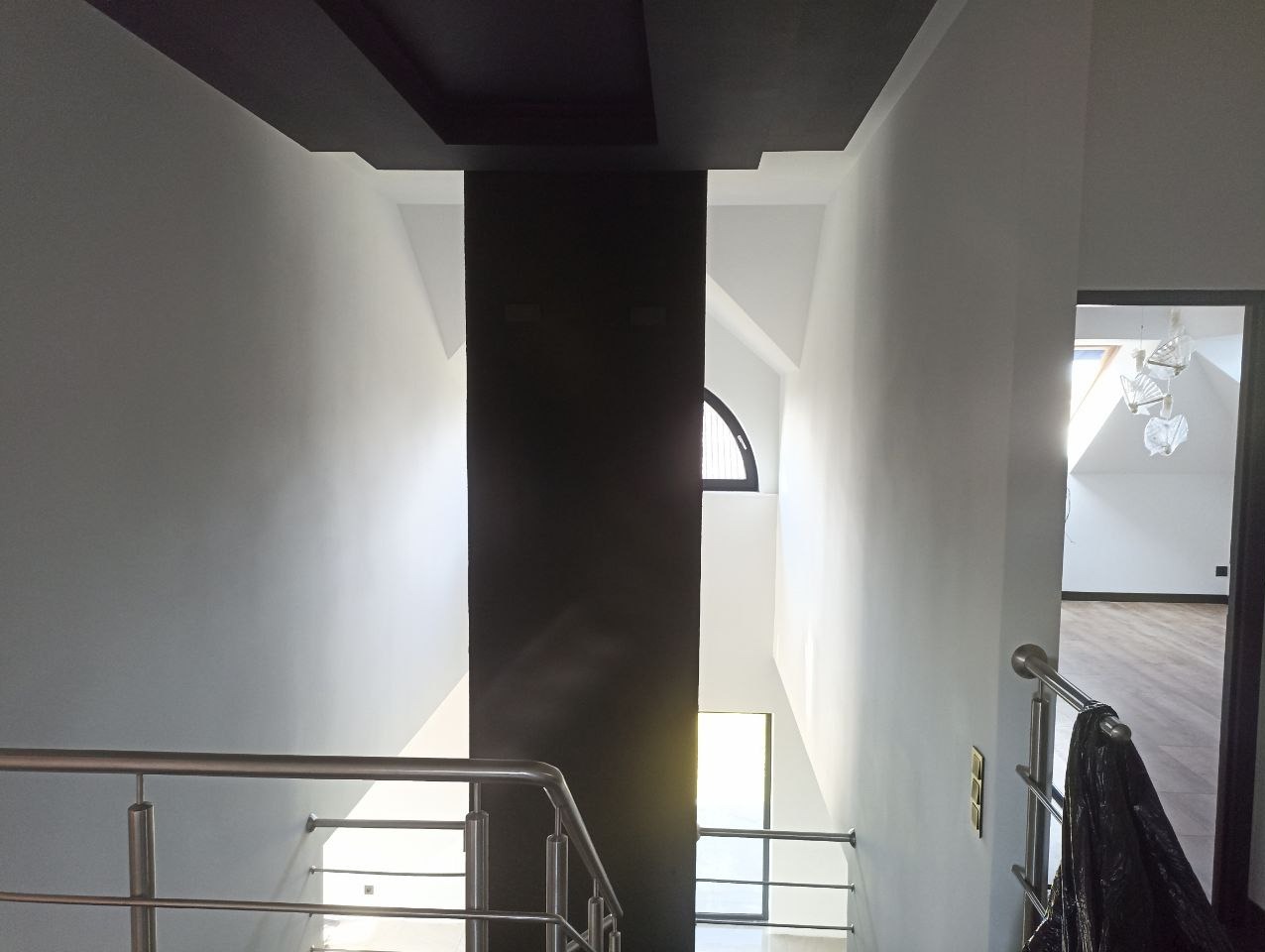 Jasne wnętrze z białymi ścianami, metalową balustradą i czarnym, centralnym elementem architektonicznym. W tle widoczne okno i designerska lampa.