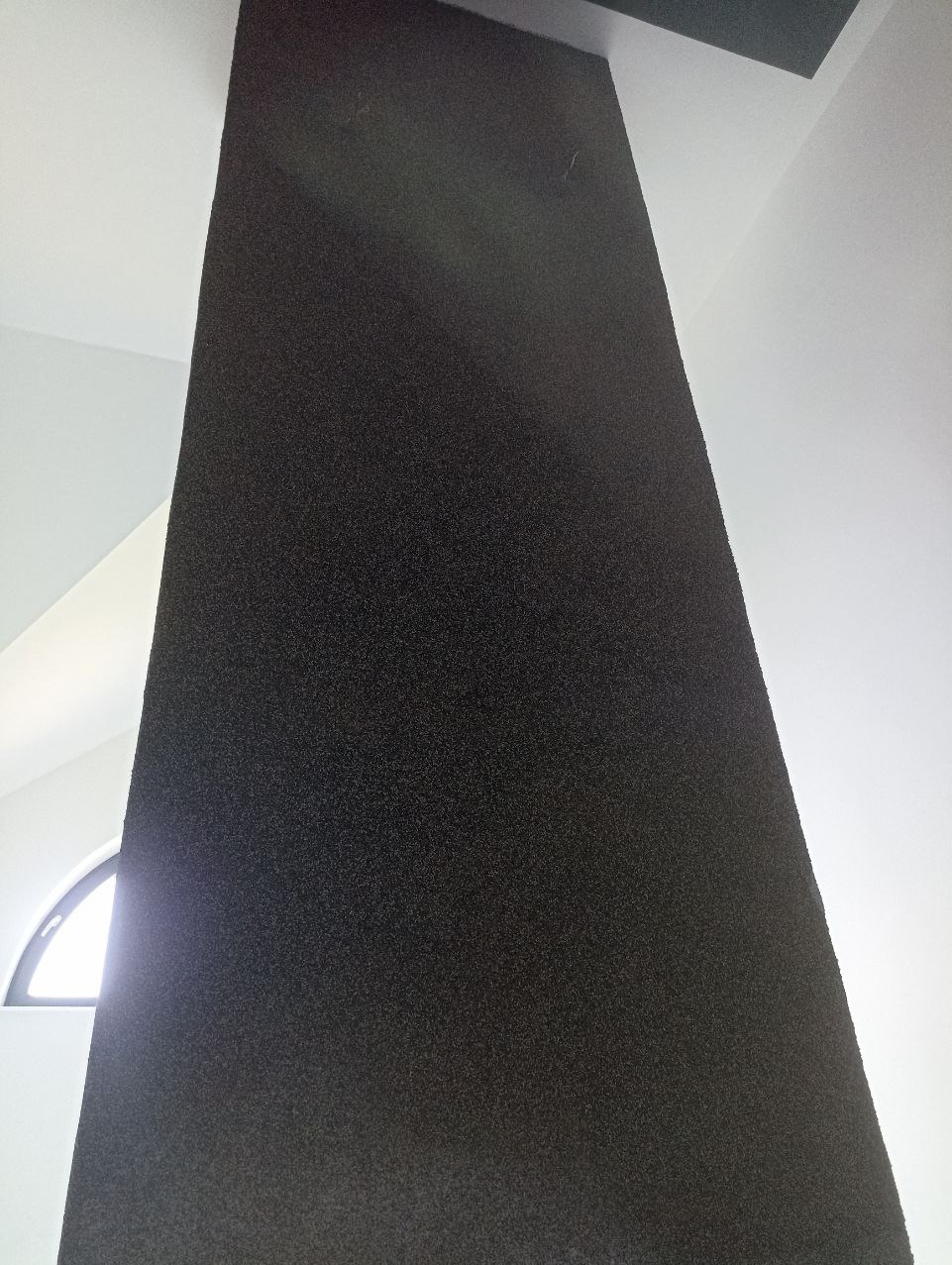 Czarny, teksturowany pionowy element architektoniczny w jasnym wnętrzu, widok z dołu, z fragmentem okna łukowego w tle. Minimalistyczny design.