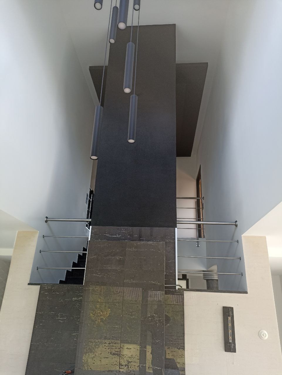 Wysoki hall z nowoczesnym oświetleniem punktowym, schodami z metalową balustradą i ciemną kolumną dekoracyjną. Minimalistyczny design.