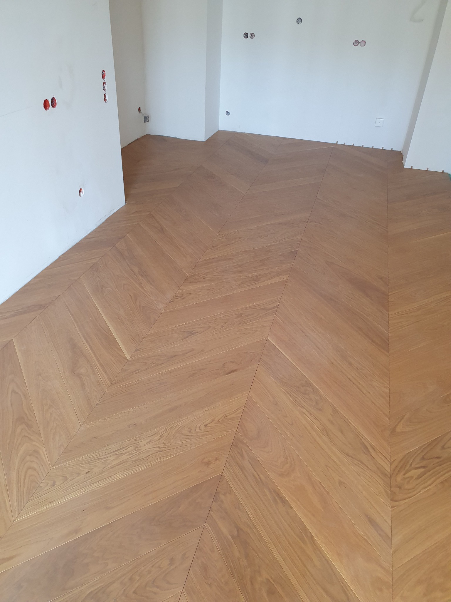 Ukończona podłoga z jasnego drewna ułożona we wzór jodełki francuskiej w pustym pomieszczeniu z białymi ścianami i widocznymi puszkami elektrycznymi.