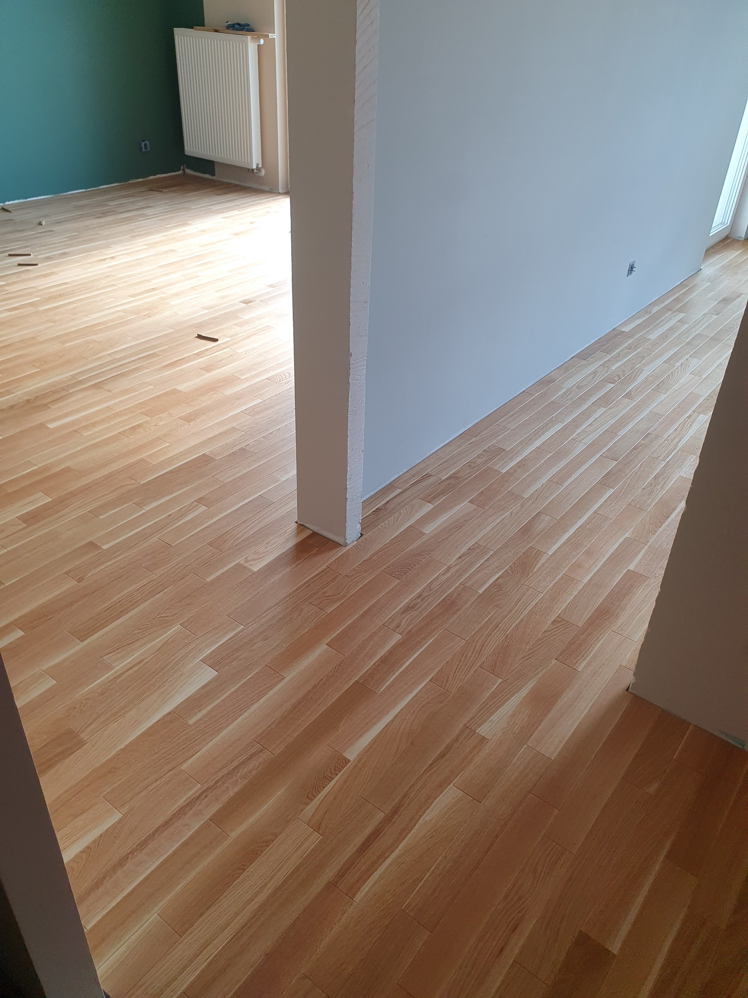 Nowo ułożony, jasny parkiet drewniany w pokoju z zieloną i szarą ścianą, widoczne niewielkie elementy montażowe na podłodze.