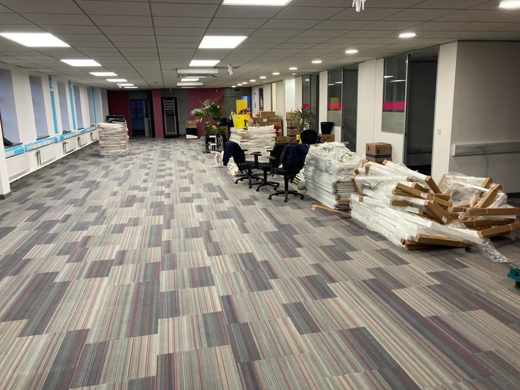 Duże biuro przed montażem mebli z nową, wzorzystą wykładziną dywanową w odcieniach szarości i czerwieni.