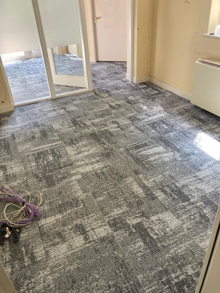 Szara wykładzina dywanowa w płytkach z abstrakcyjnym wzorem w biurze, widoczne kable elektryczne w rogu pomieszczenia.