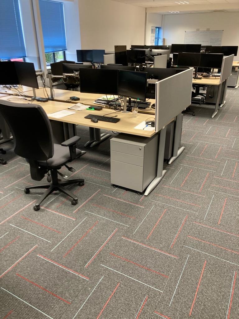 Biuro z szarą wykładziną dywanową w geometryczny wzór, biurkami z przegrodami i czarnymi monitorami.