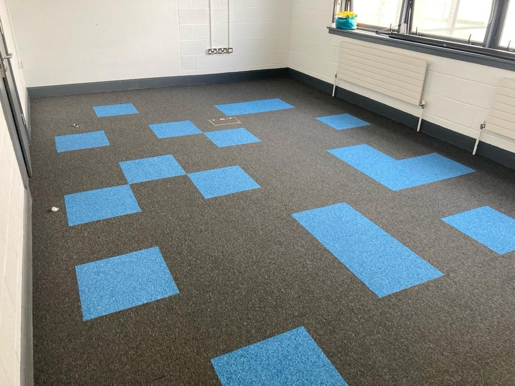 Wykładzina dywanowa w odcieniach szarości z niebieskimi, geometrycznymi wstawkami w kształcie kwadratów i prostokątów, ułożona w jasnym pomieszczeniu z grzejnikami pod oknem.