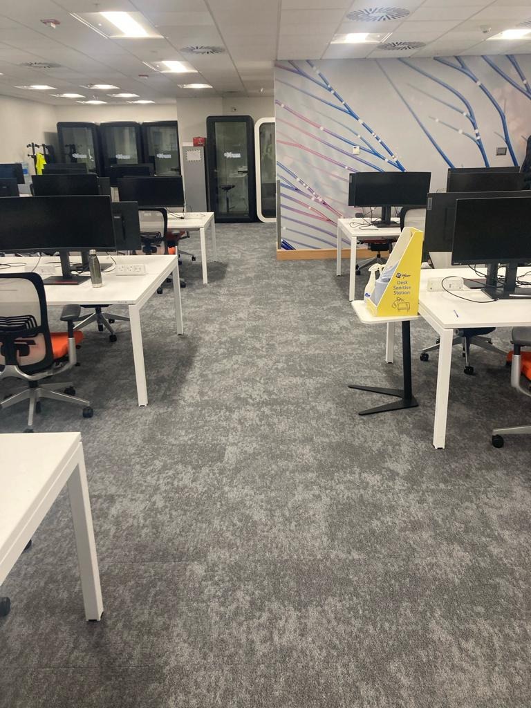 Biuro z szarą wykładziną dywanową w odcieniach szarości, białe biurka z komputerami, kabiny telefoniczne w tle.