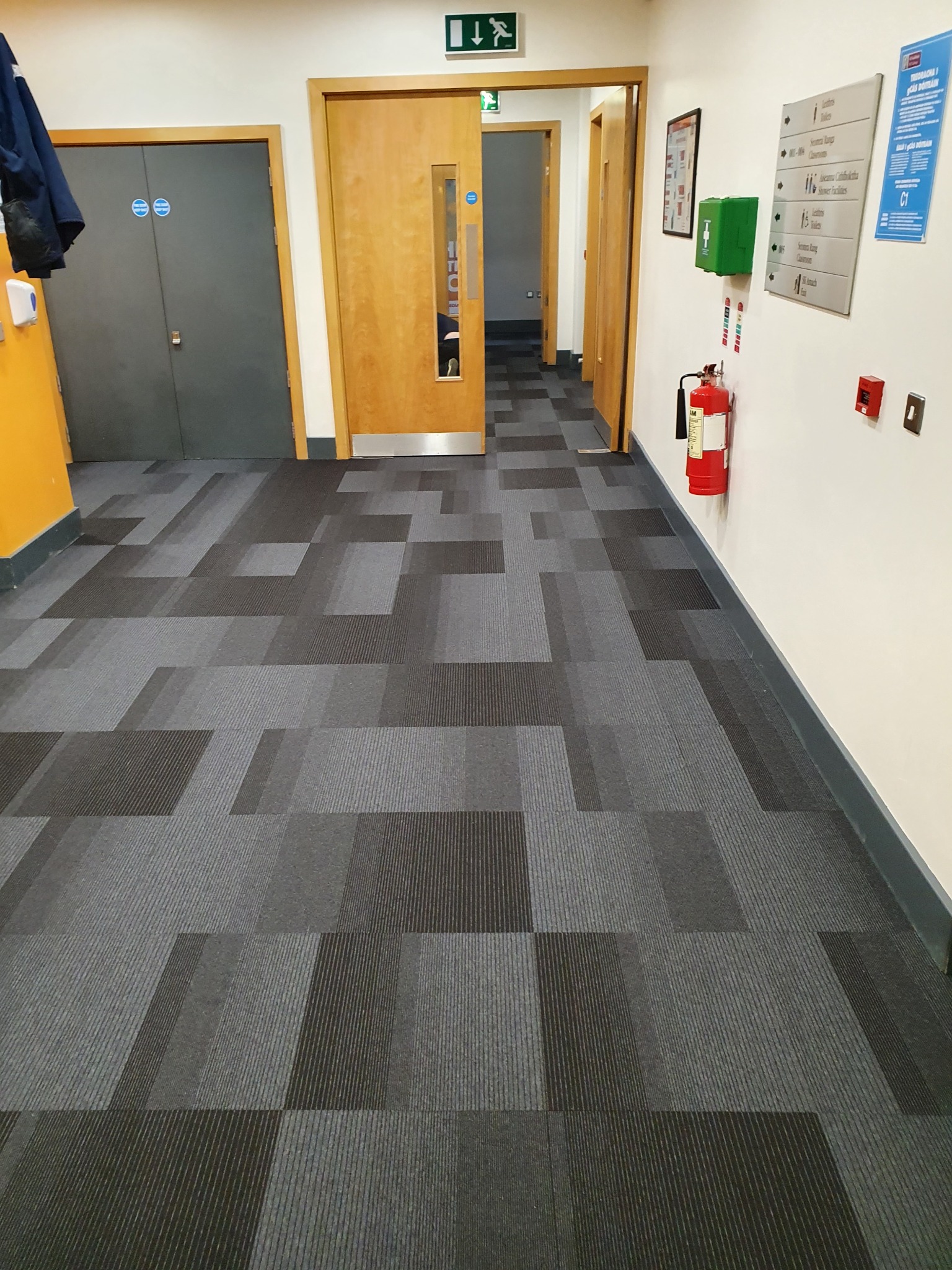 Wzór geometryczny z szarych i czarnych prostokątów na podłodze korytarza, widoczne drzwi wejściowe i znaki informacyjne na ścianie.